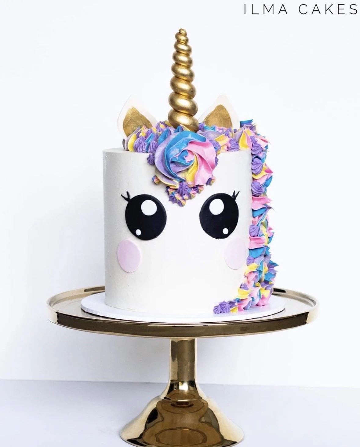 Unicorn-themed birthday cake with white icing, large black and white cartoon eyes, pink cheeks, a golden horn, and colorful rainbow frosting on the mane, displayed on a gold cake stand. The top right corner has the text 'ILMA CAKES'.