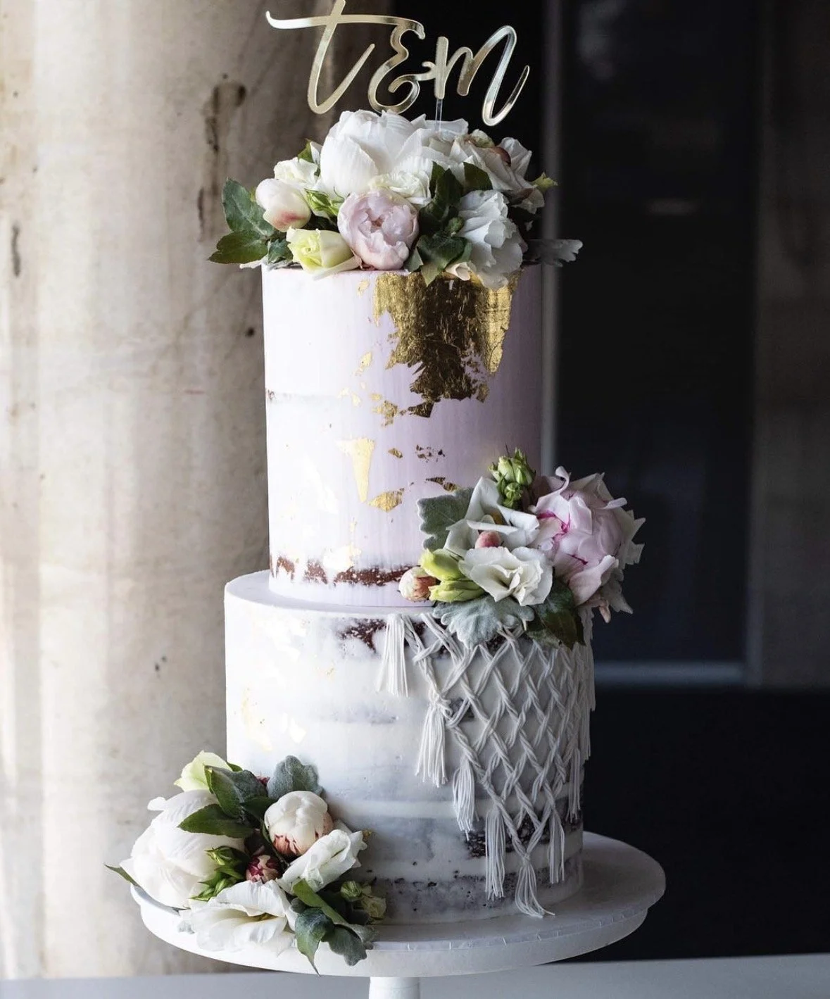 Elegant three-tier wedding cake with gold leaf accents and floral decorations, topped with a "t&M" cake topper.