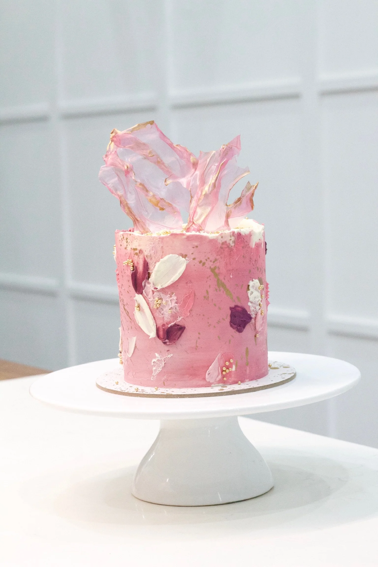 A pink, cylindrical cake decorated with white, purple, and gold accents sits on a white cake stand. The top features pink and gold sugar decorations.
