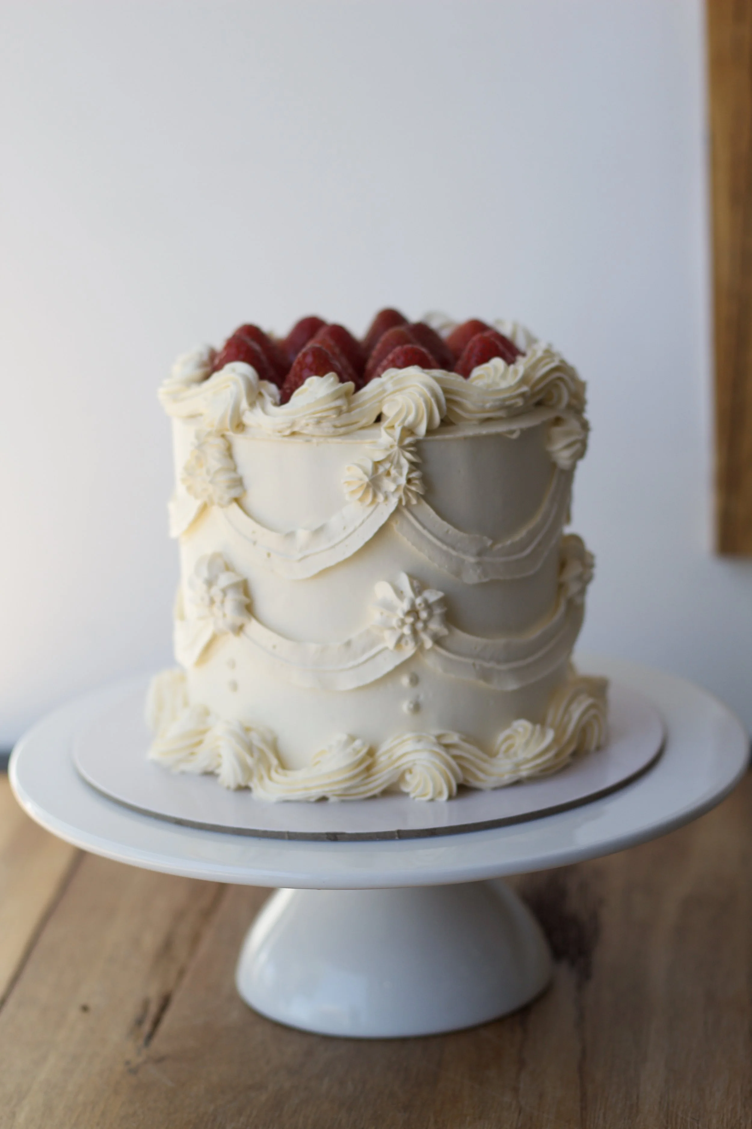 A stunning retro multi-tiered white cake decorated with cream-colored frosting and fresh strawberries on top, displayed on a white cake stand available online through the online ordering system with 3 business days notice 