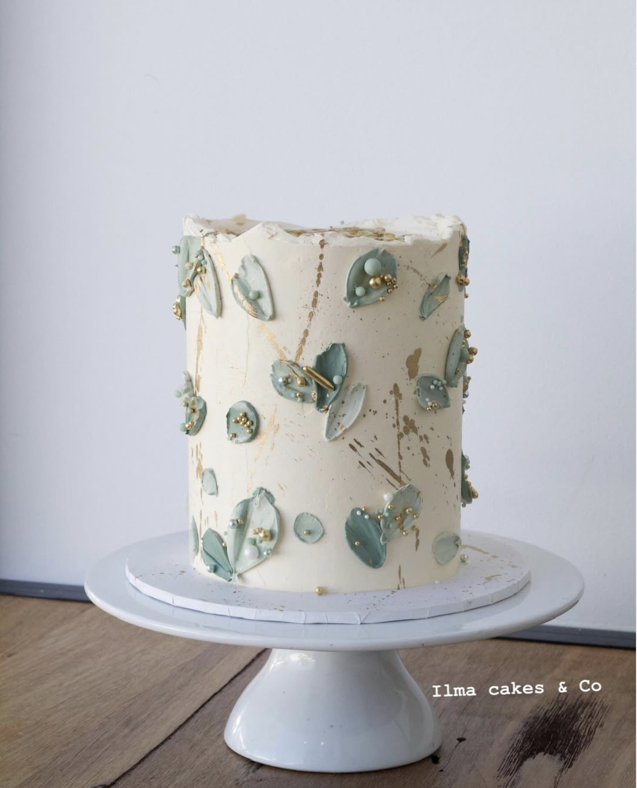 A tall white cake decorated with greenish-blue leaves and gold pearls on a white cake stand.