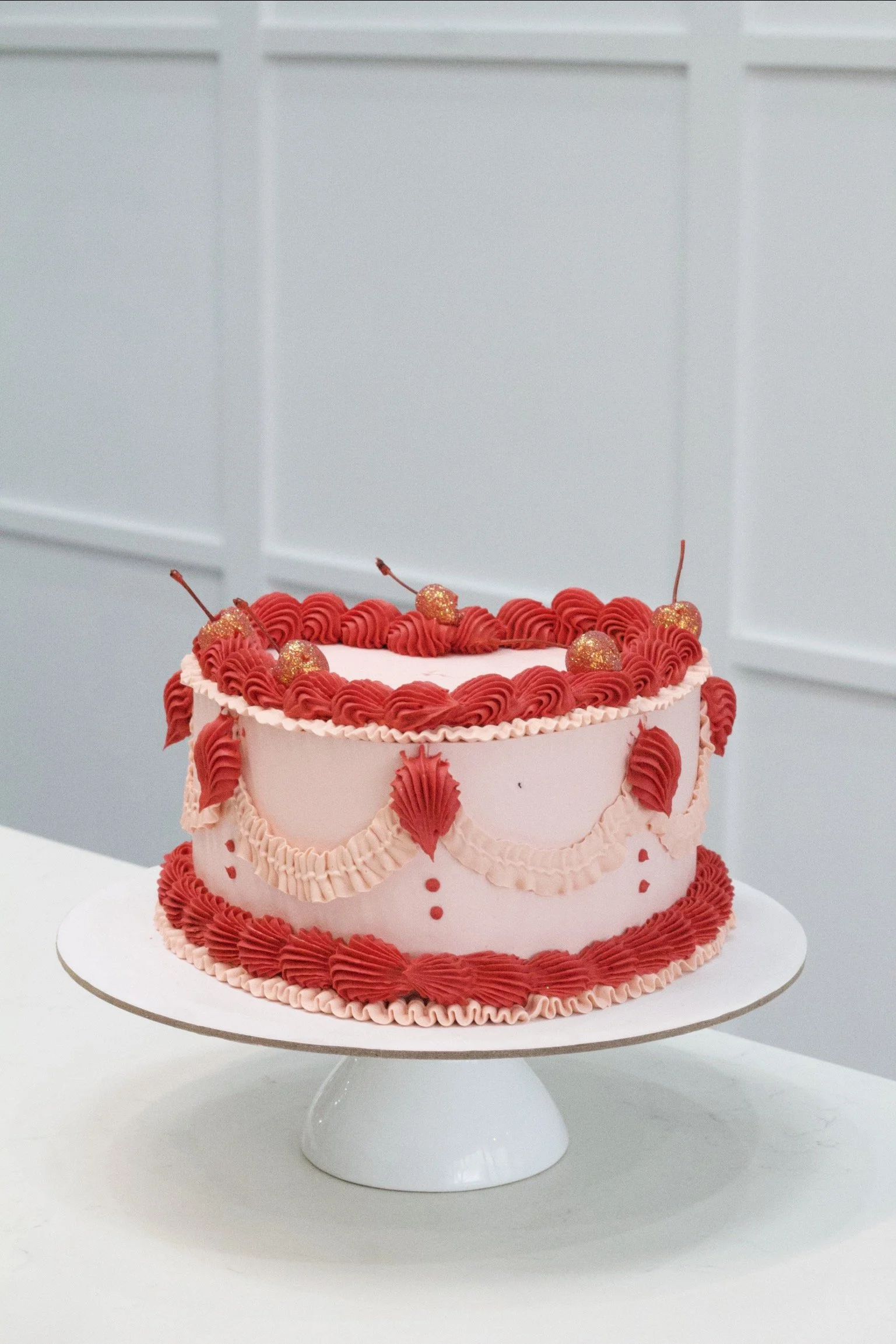A pink cake decorated with red and pink frosting, small gold cherries on top, and pink ruffles around the edges, displayed on a white cake stand.