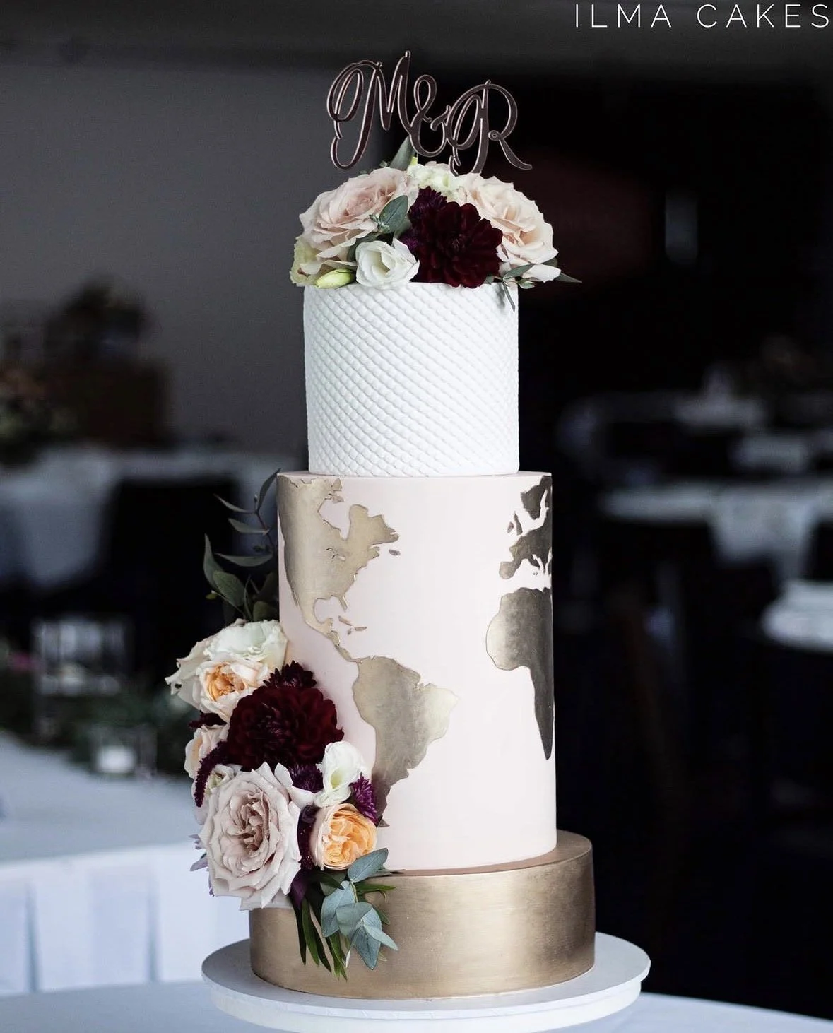 A three-tier wedding cake with floral decorations and a world map design. The top tier is white with a textured pattern and topped with fresh flowers and a "Mr & Mrs" cake topper. The middle tier has a cream-colored background with gold world map out