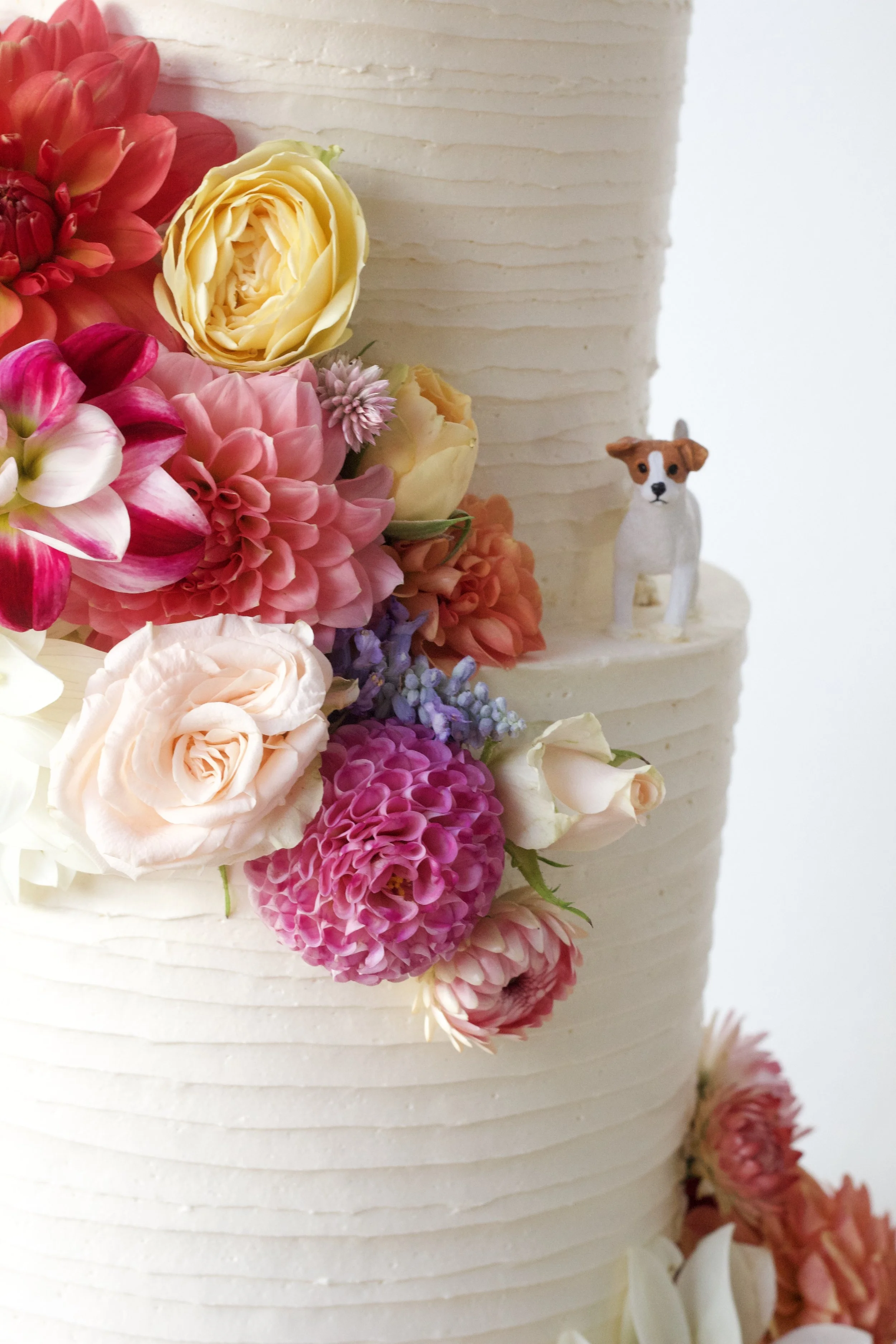 A close-up of a white cake decorated with colorful pink, yellow, and purple flowers, and a small figurine of a dog on the side.