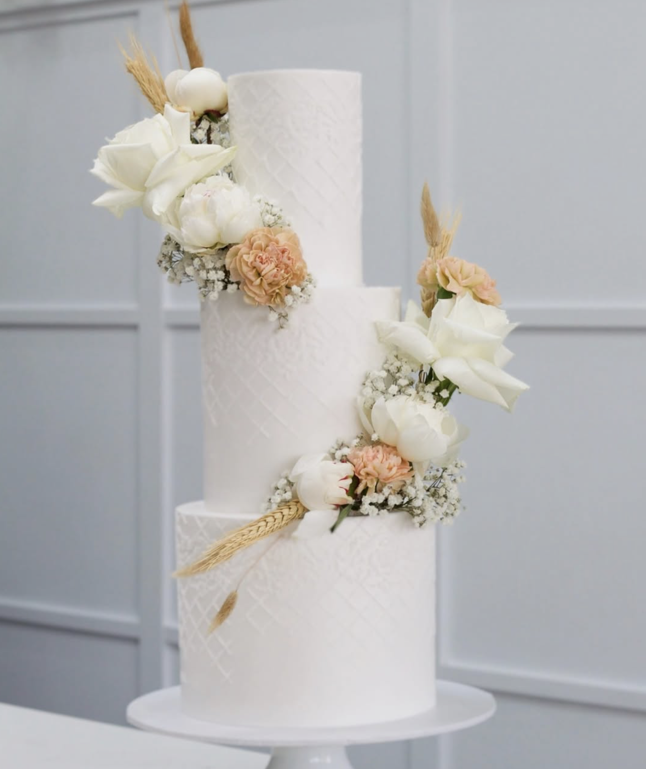 A three-tier white wedding cake decorated with white roses, peach carnation flowers, baby's breath, and wheat stalks.