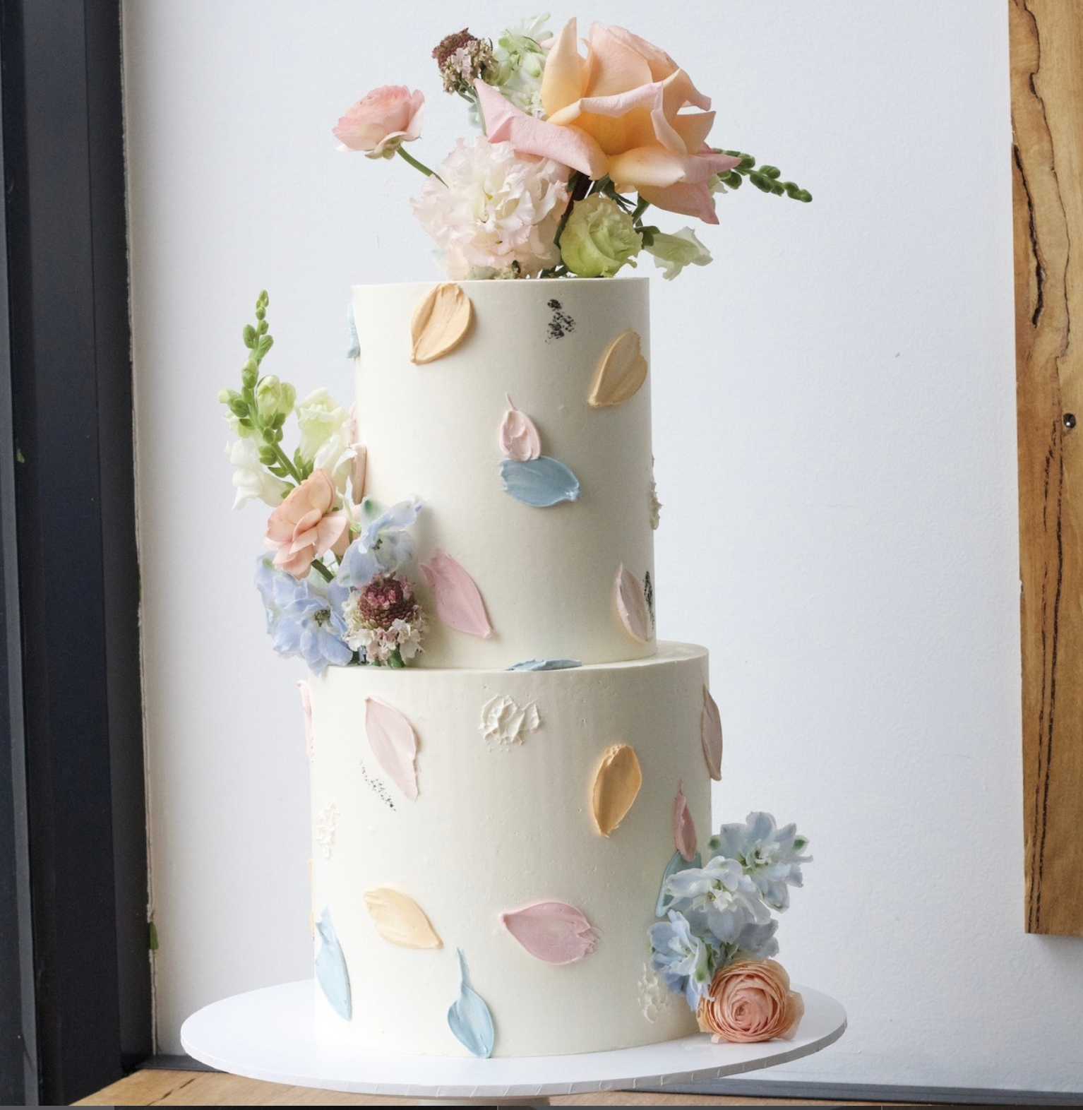 A three-tiered wedding cake decorated with pastel-colored flower petals and fresh flowers, including roses and hydrangeas, on a white cake stand against a plain white background.