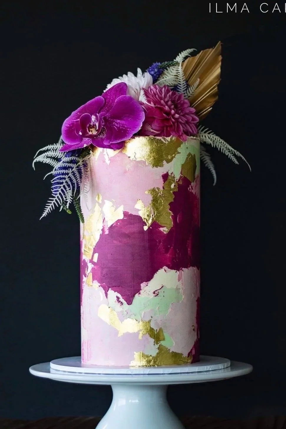 A tall, cylindrical cake with pink, gold, and white marbled design, decorated  exceptionally with purple orchids, pink flowers, white and purple foliage, and a gold leaf accent. Custom cakes are their specialty