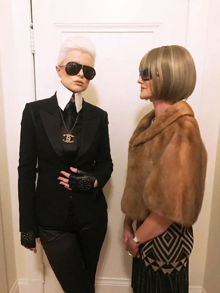 Two women dressed in vintage glamorous style, both wearing sunglasses. The woman on the left has short white hair, a black suit with a white shirt and black tie, and wears a Chanel necklace. The woman on the right has a bob haircut, a fur shawl, and 