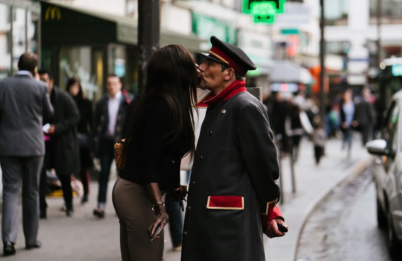 A man and woman kiss on a busy city street surrounded by pedestrians and vehicles.