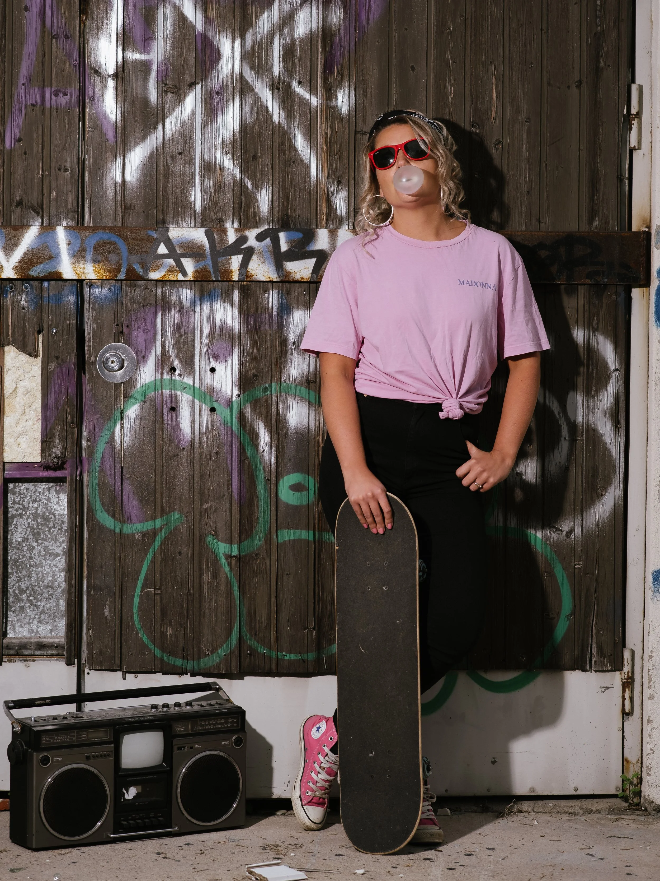 A woman wearing red sunglasses and a pink Madonna t-shirt blows a bubble gum bubble while leaning on a skateboard. She is standing in front of a graffiti-covered wooden wall and a vintage stereo.