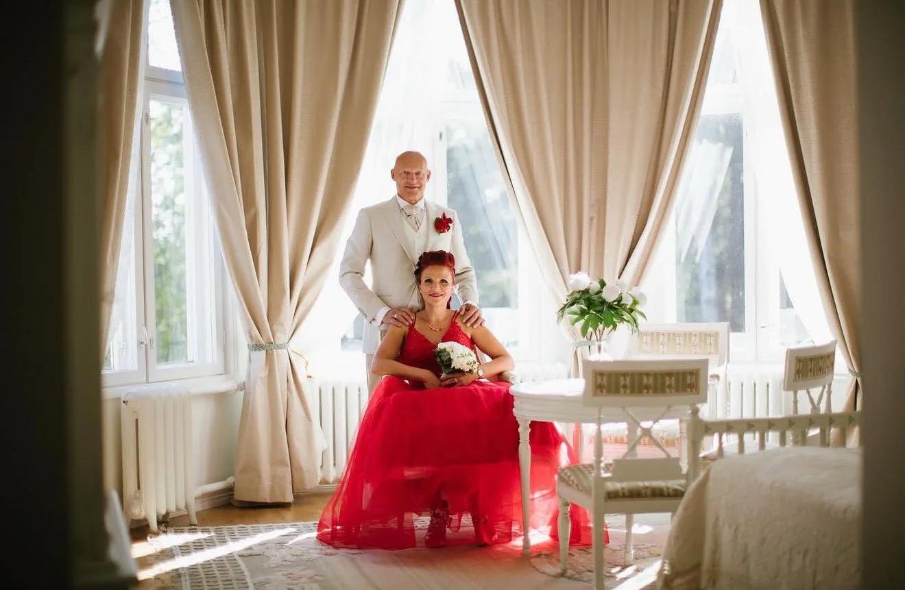 A couple dressed in wedding attire, with the woman seated and holding a bouquet and the man standing behind her, in a bright room with large windows and cream curtains.