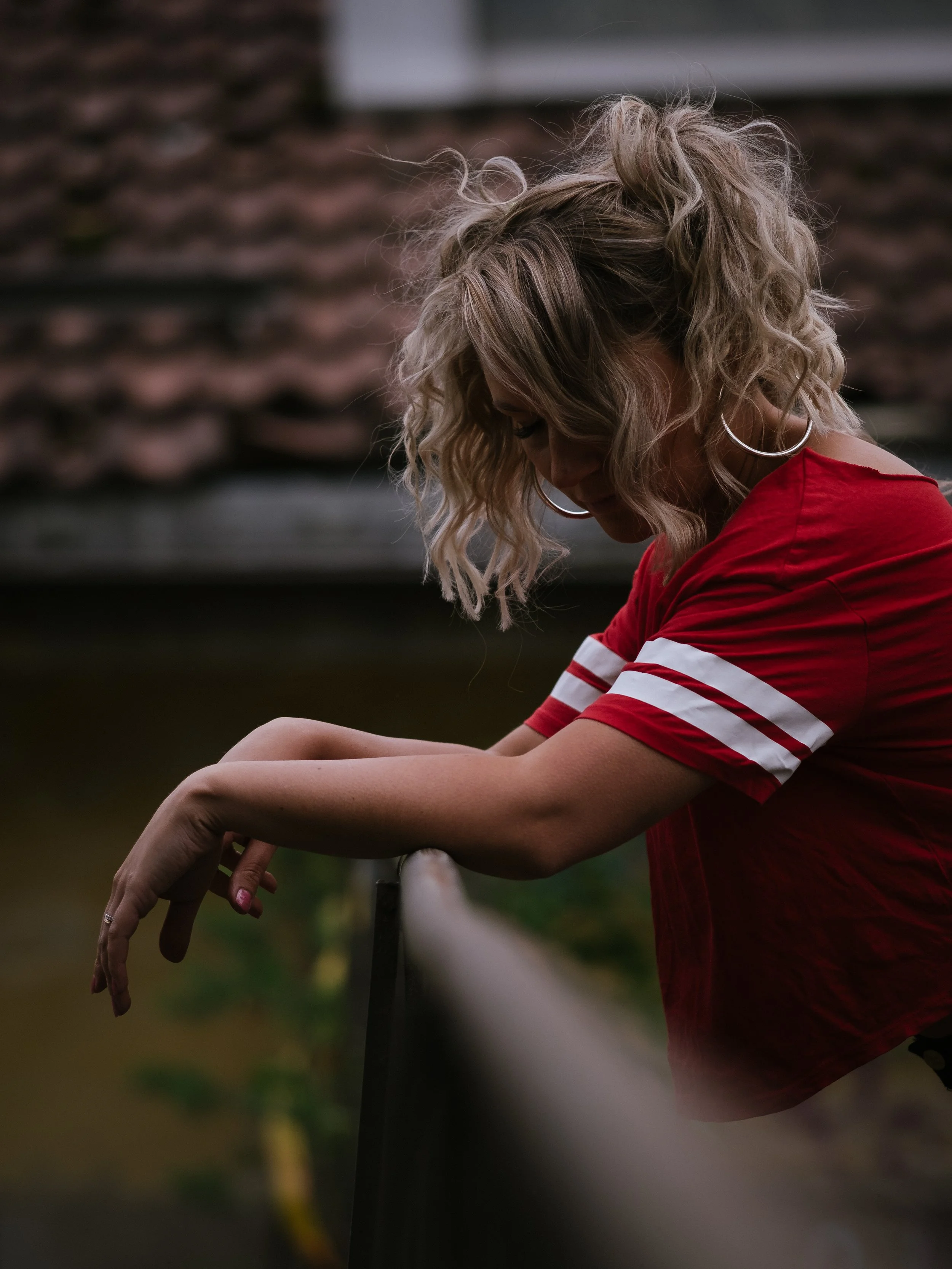 A woman with curly blonde hair wearing a red shirt with white stripes on the sleeves, leaning on a fence with her arms crossed, looking down.