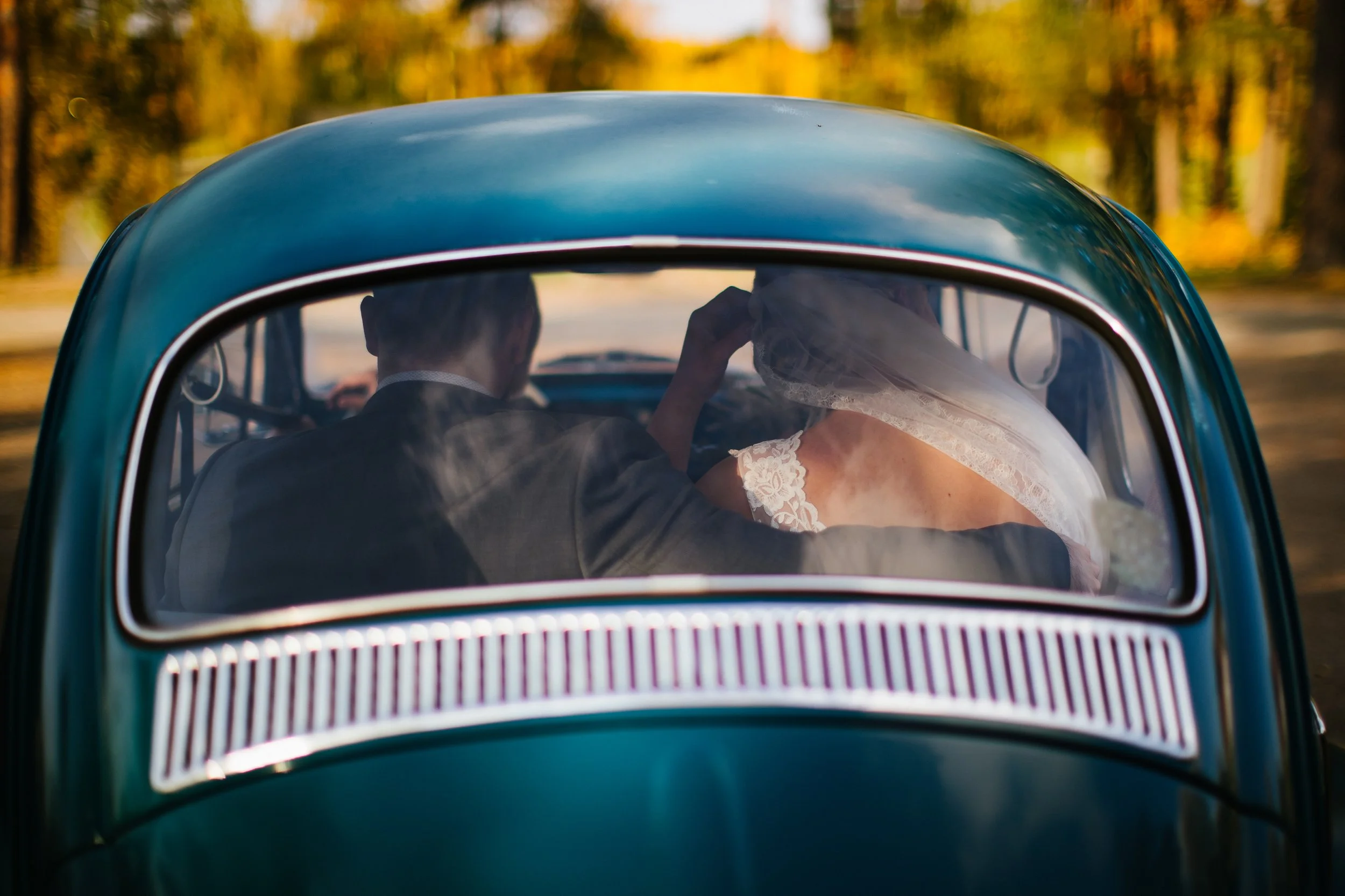 The image shows a vintage car with a blue exterior, viewed from the rear window. Inside the car, a bride wearing a wedding dress and veil, and a groom in a dark suit, are sitting close together, with the bride touching her face, possibly adjusting he
