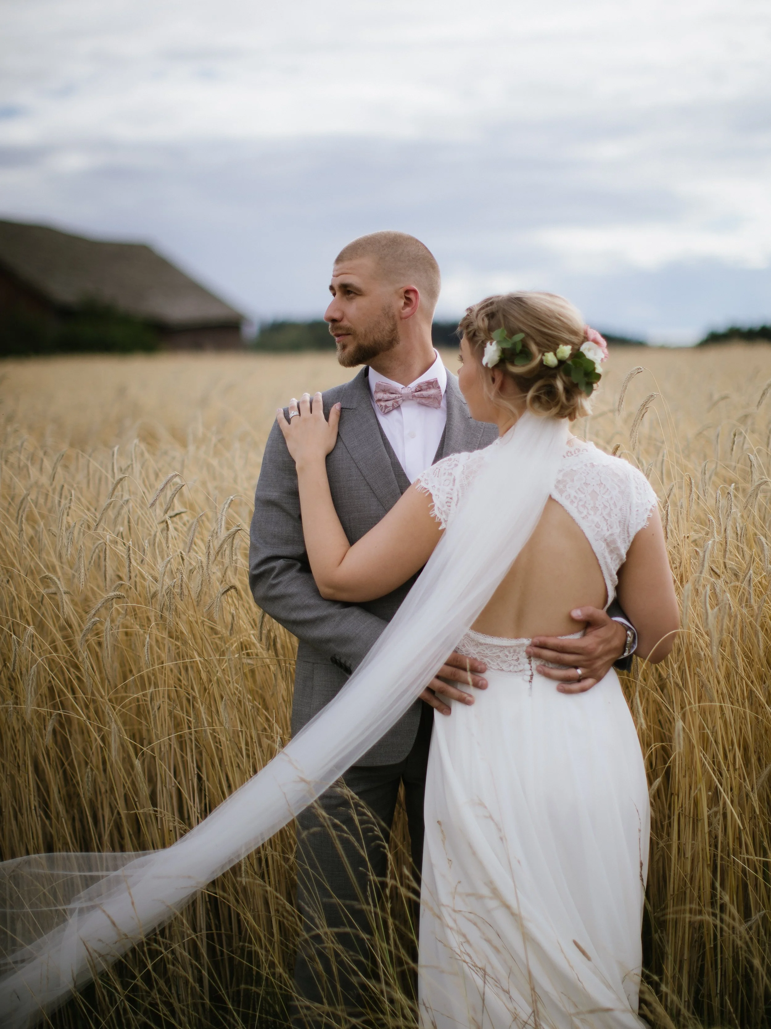 A bride and groom embrace in a wheat field with a barn in the background, under cloudy skies.