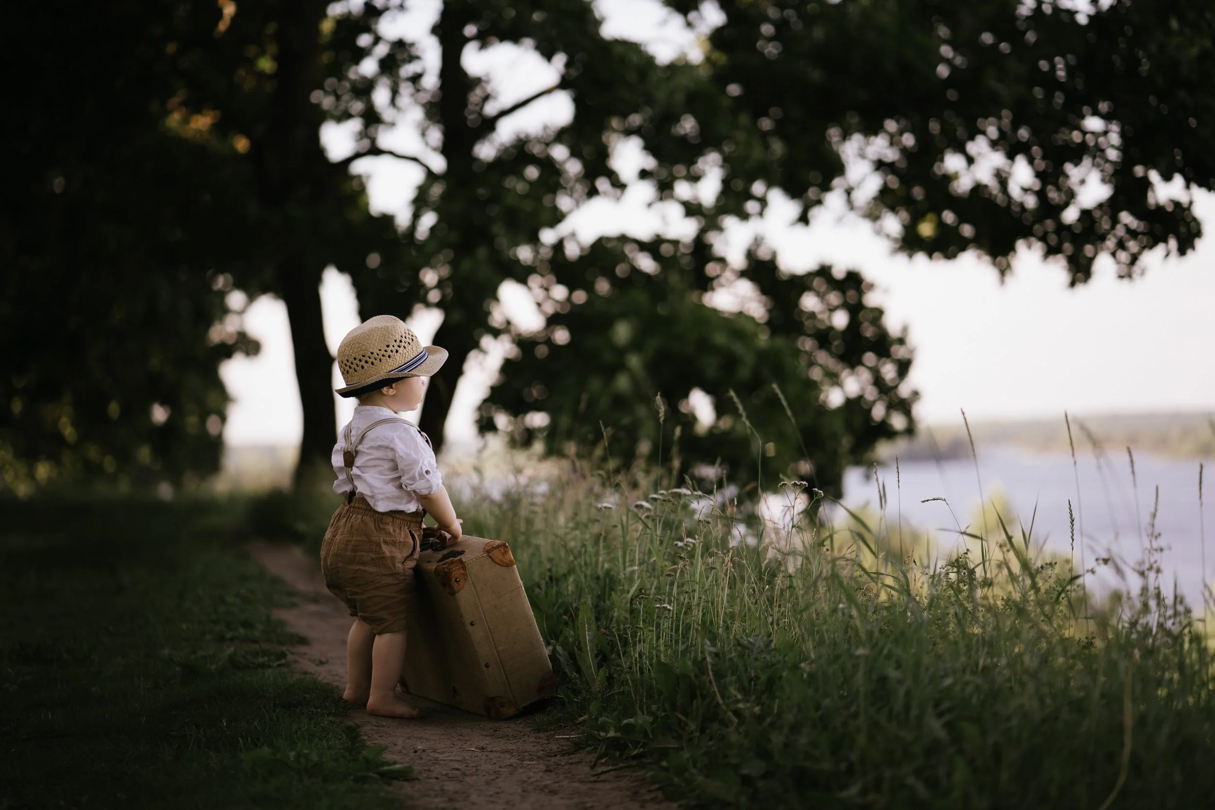 A young child with a straw hat and suspenders stands by a wooden suitcase on a dirt path, overlooking a body of water and trees in the background during sunset or sunrise.