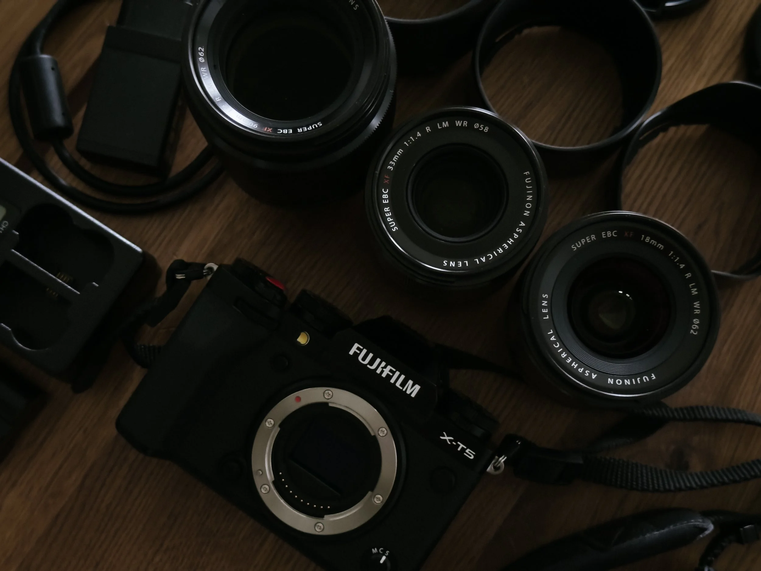 Various camera lenses and camera accessories scattered on a wooden surface, including multiple camera lenses, lens caps, and a camera body.