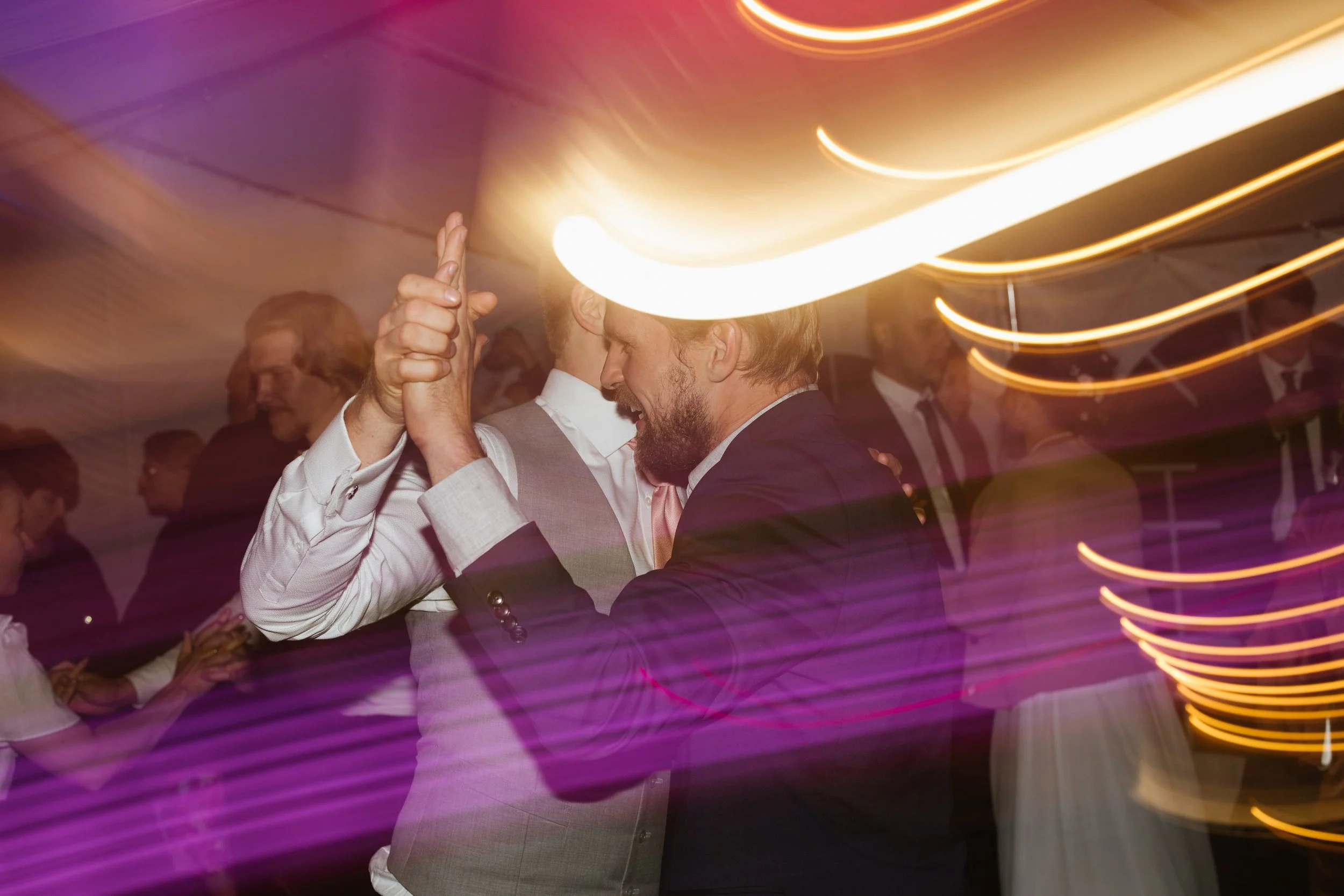 Two men in formal attire dancing and holding hands at a party or wedding reception, surrounded by other guests and colorful light streaks.