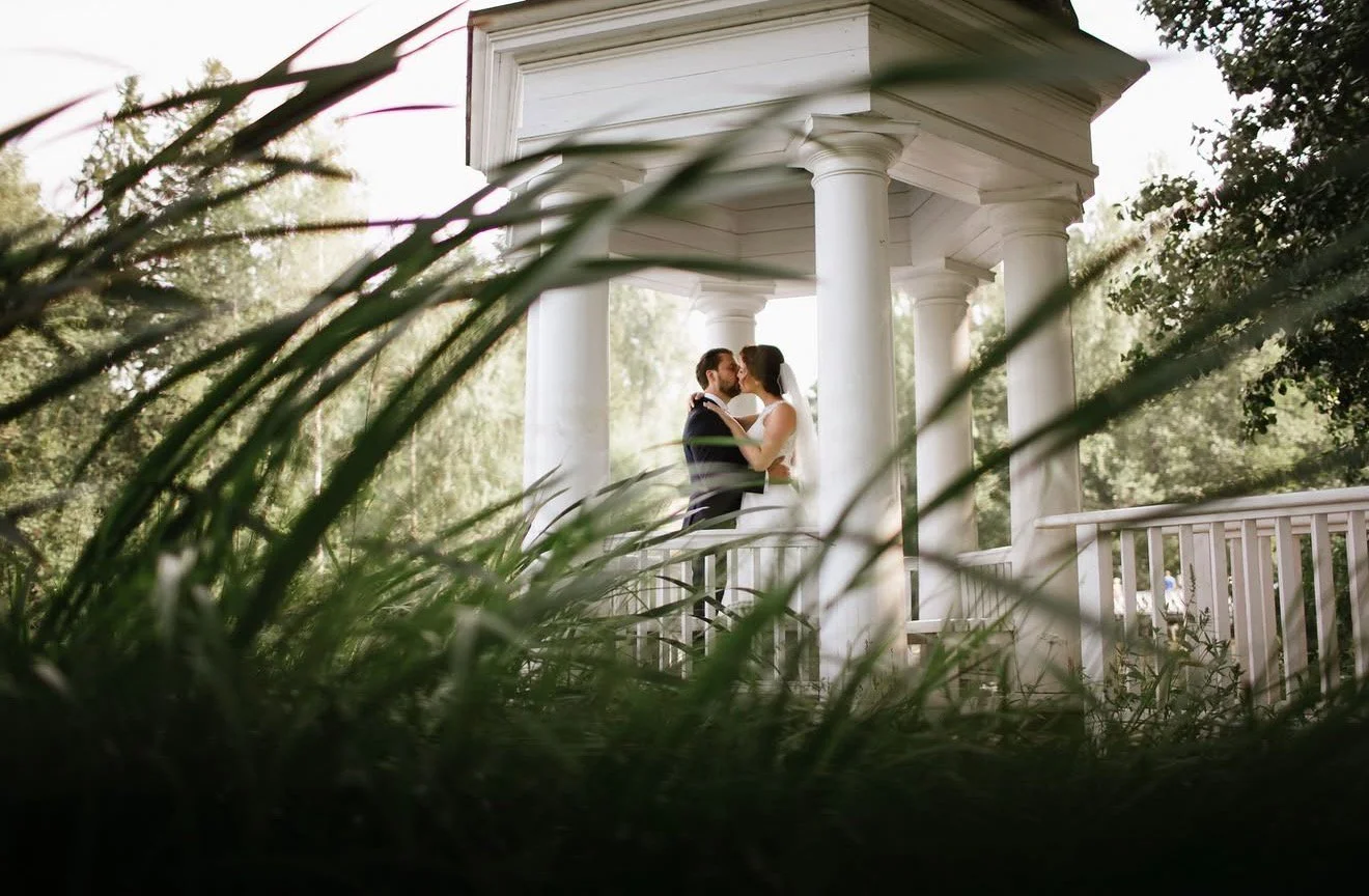 A bride and groom sharing a kiss on a white pavilion surrounded by greenery.