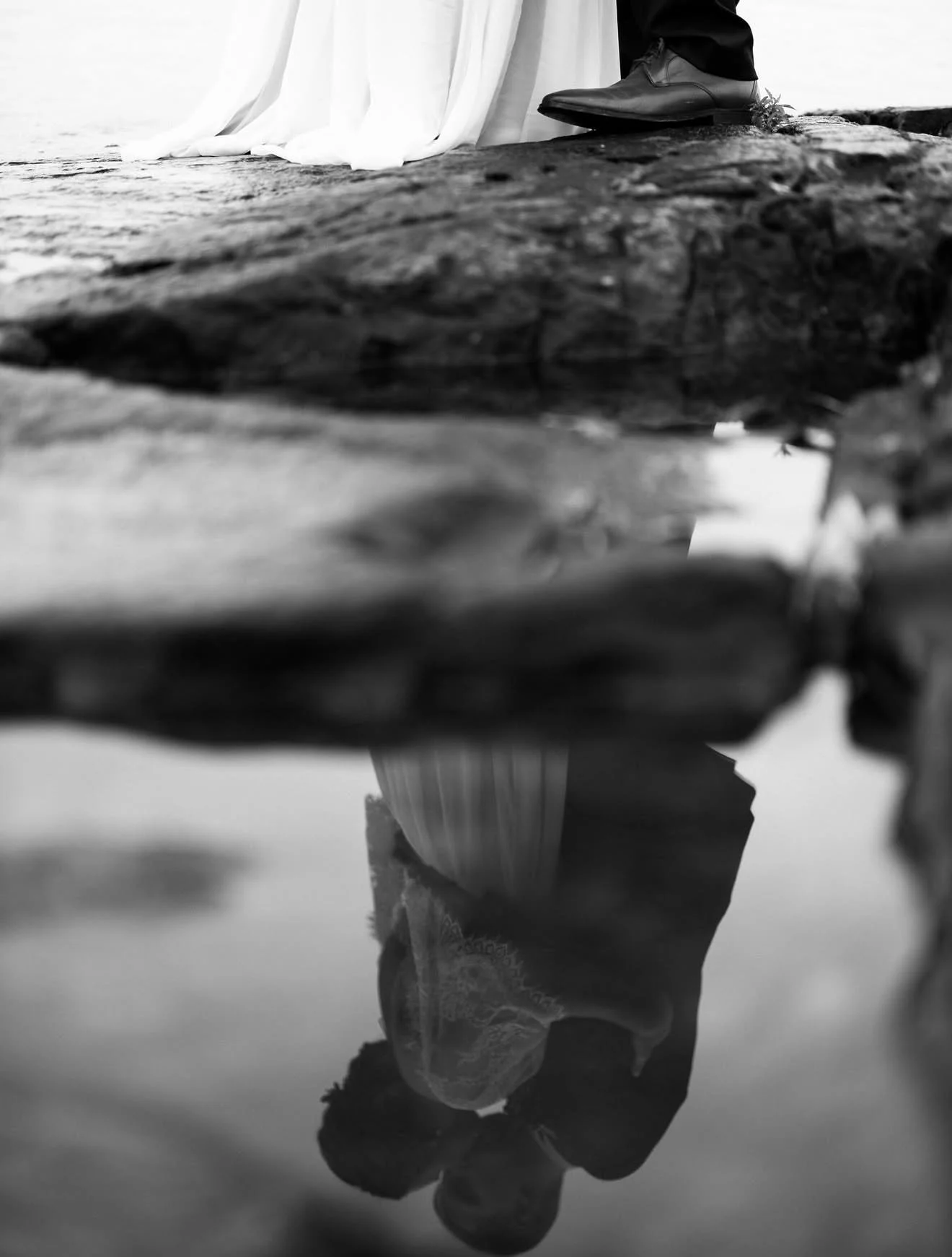 A black and white photo of a person standing near water, with their reflection visible on the surface.