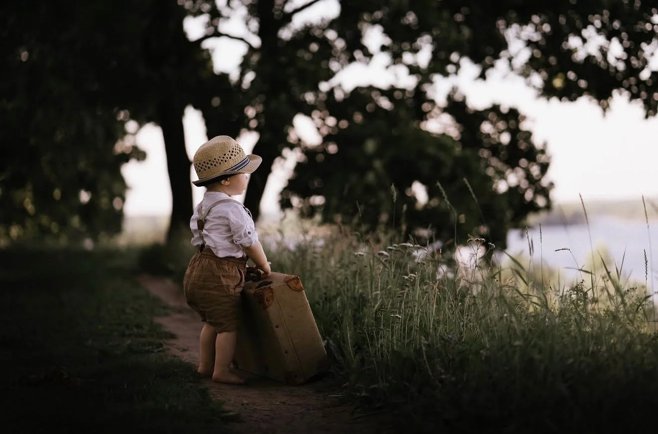 A young child wearing a straw hat and vintage clothing, standing barefoot on a dirt path near tall grass, looking at a body of water in the distance, surrounded by trees, during what appears to be late afternoon or early evening.