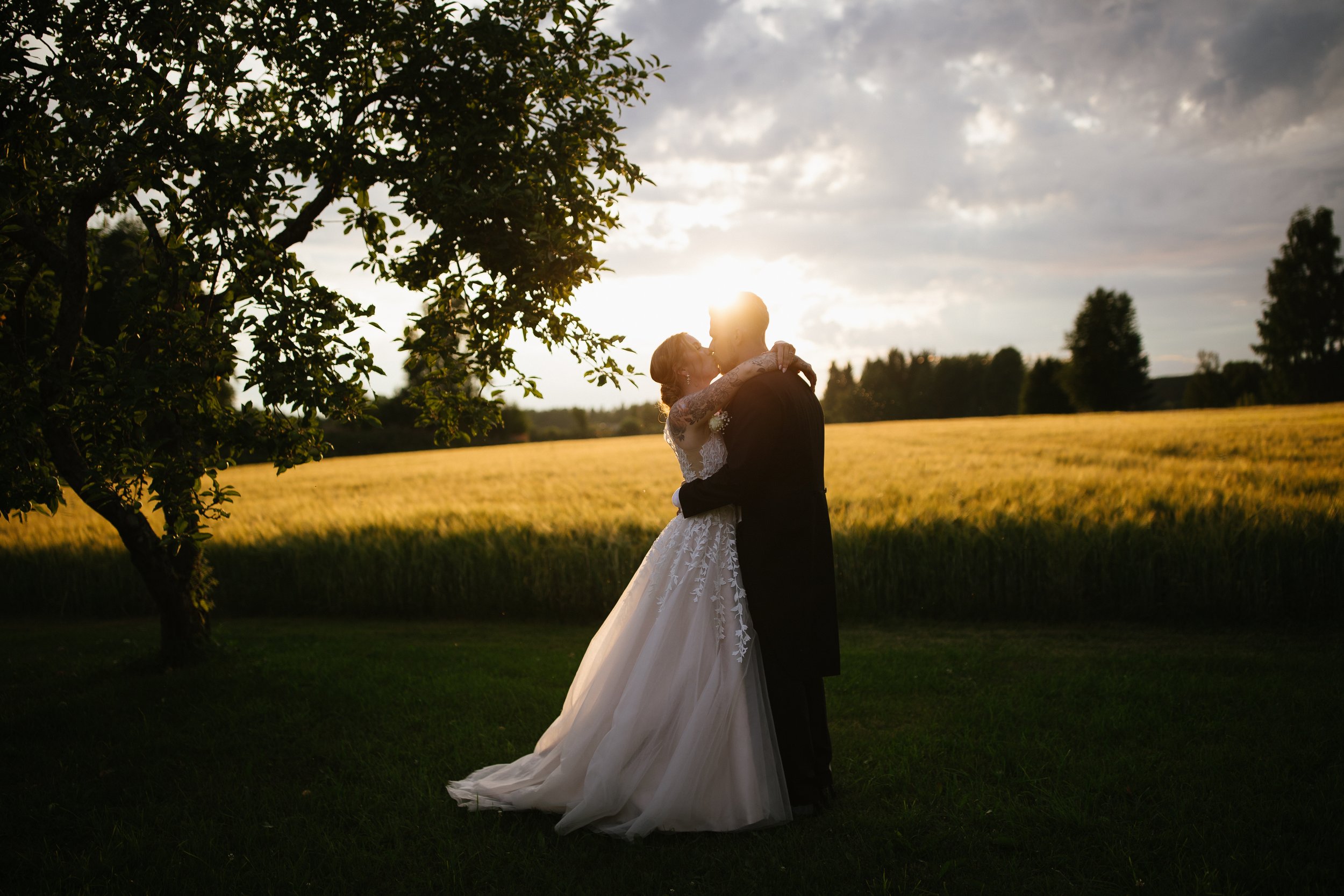 A bride and groom embrace and kiss outdoors during sunset, standing on a grassy field with trees and a cloudy sky in the background.