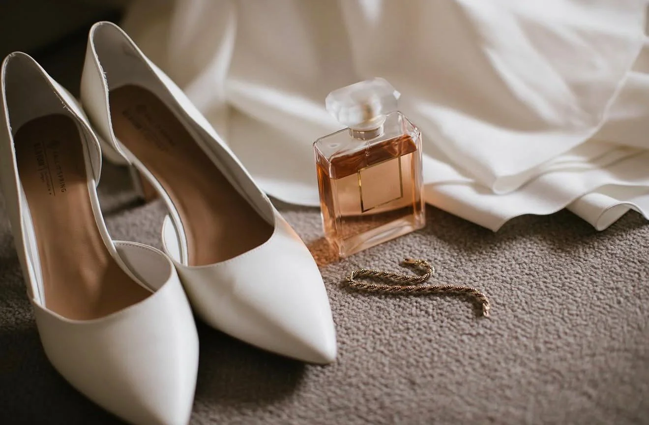 A pair of white high-heeled shoes, a bottle of perfume, a gold necklace, and a white fabric, possibly a dress, are arranged on a beige carpeted floor.