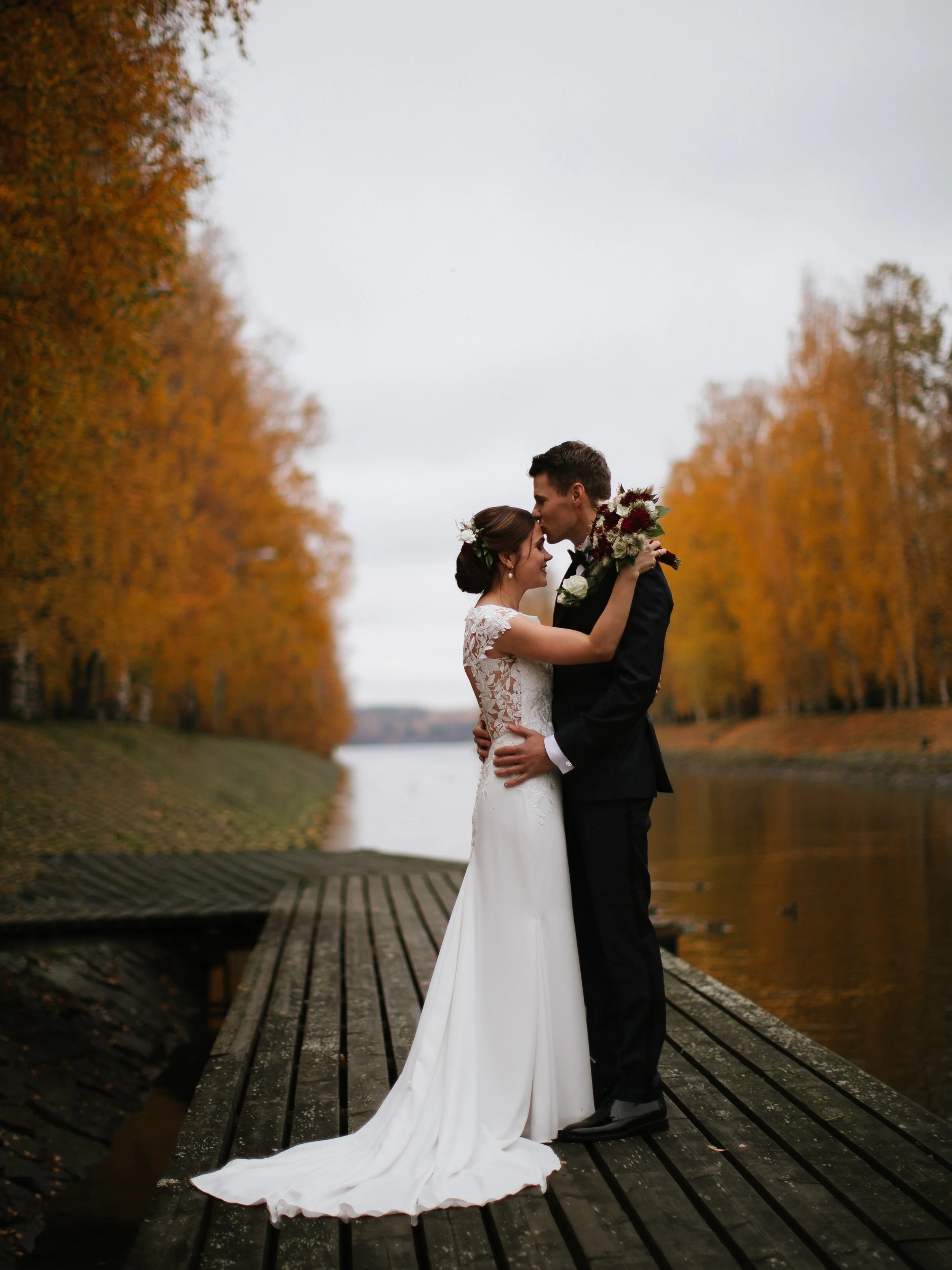 A bride and groom standing close together on a wooden dock by a river, surrounded by tall, orange autumn trees, with the groom kissing the bride's forehead.