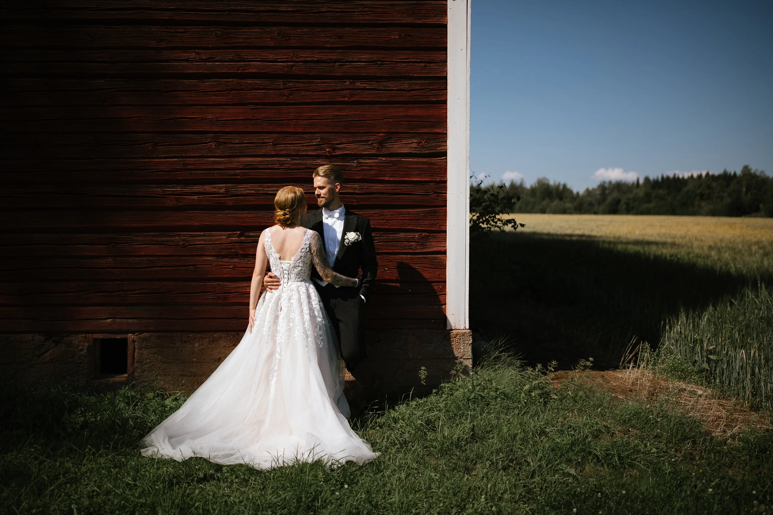 Bride and groom standing close together outside near a wooden building, with a field and trees in the background, under a clear blue sky.