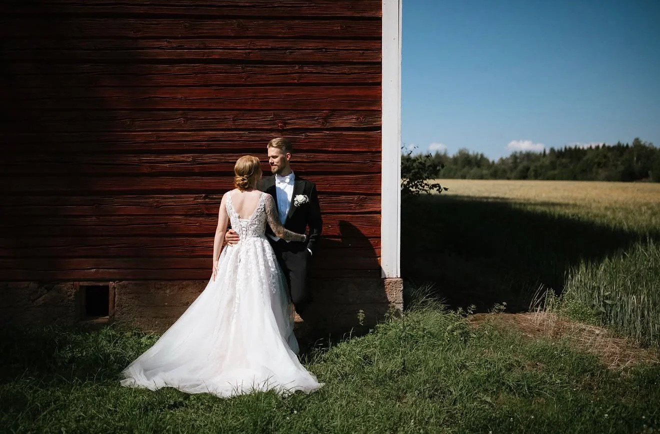 A bride and groom standing close together outdoors near a wooden building, with the bride in a white wedding dress and the groom in a black suit, on a sunny day.