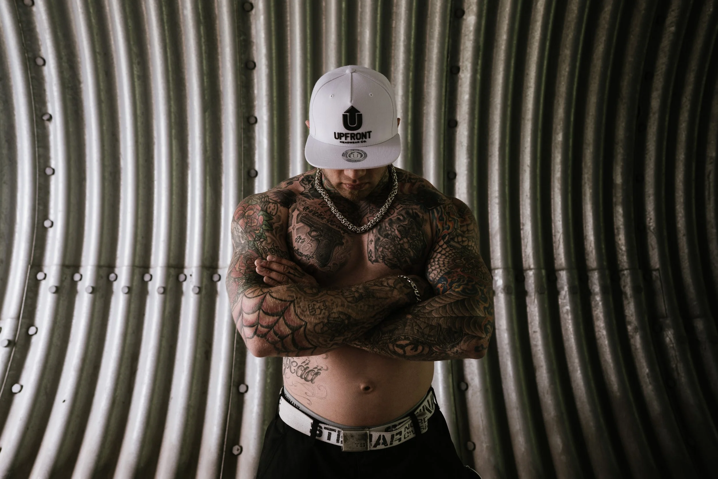 A shirtless man with tattoos covering his arms, chest, and abdomen, wearing a white cap with the logo 'UPFRONT HEADWEAR CO.' and a silver chain necklace, standing with arms crossed in front of a silver metallic corrugated wall.