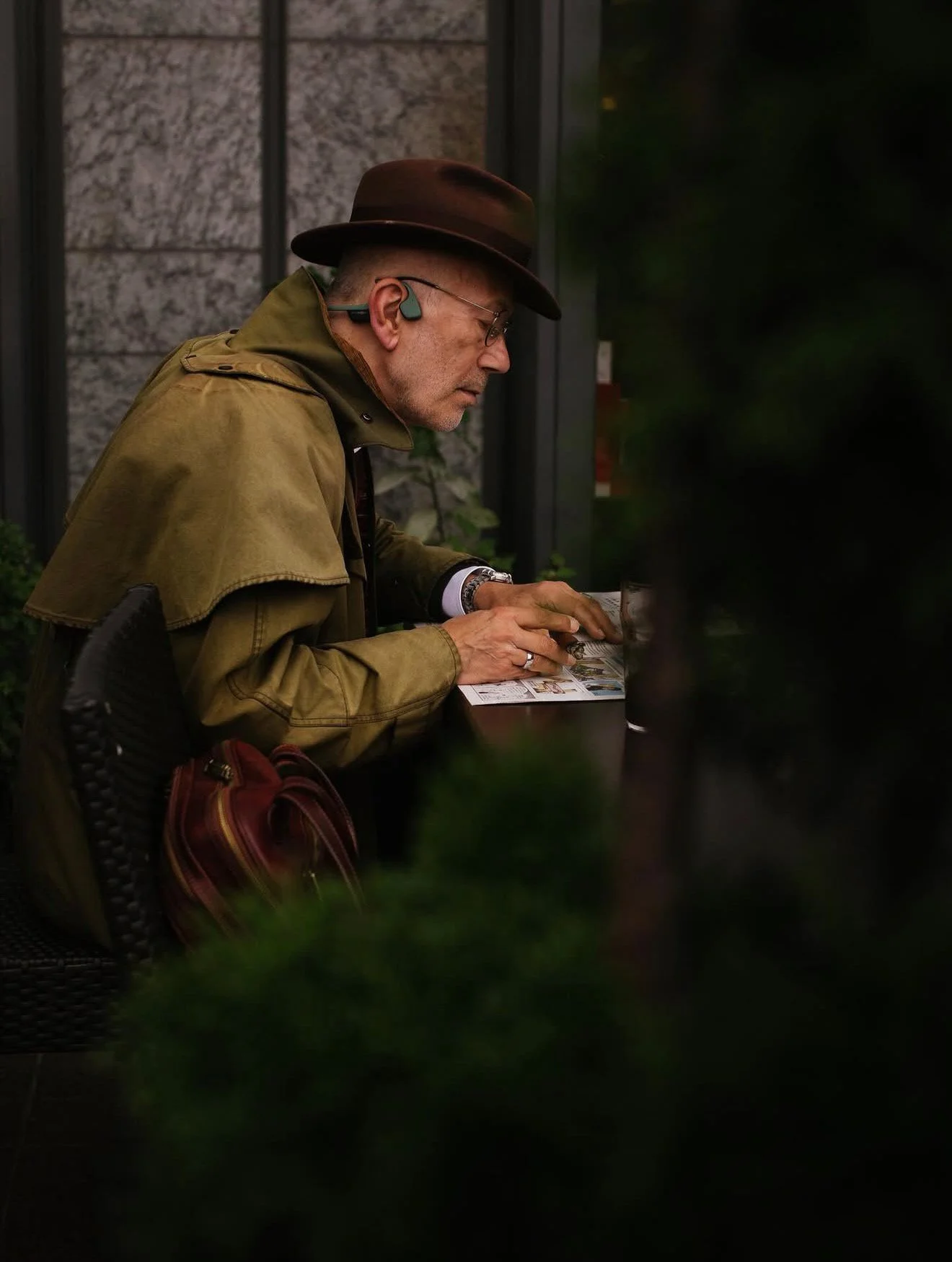 A man wearing a brown hat, khaki jacket, and glasses is sitting at a table reading a brochure or magazine. He has hearing aids and a watch on his left wrist, and a red bag is on the back of his chair. The background shows a grayish stone wall and a g