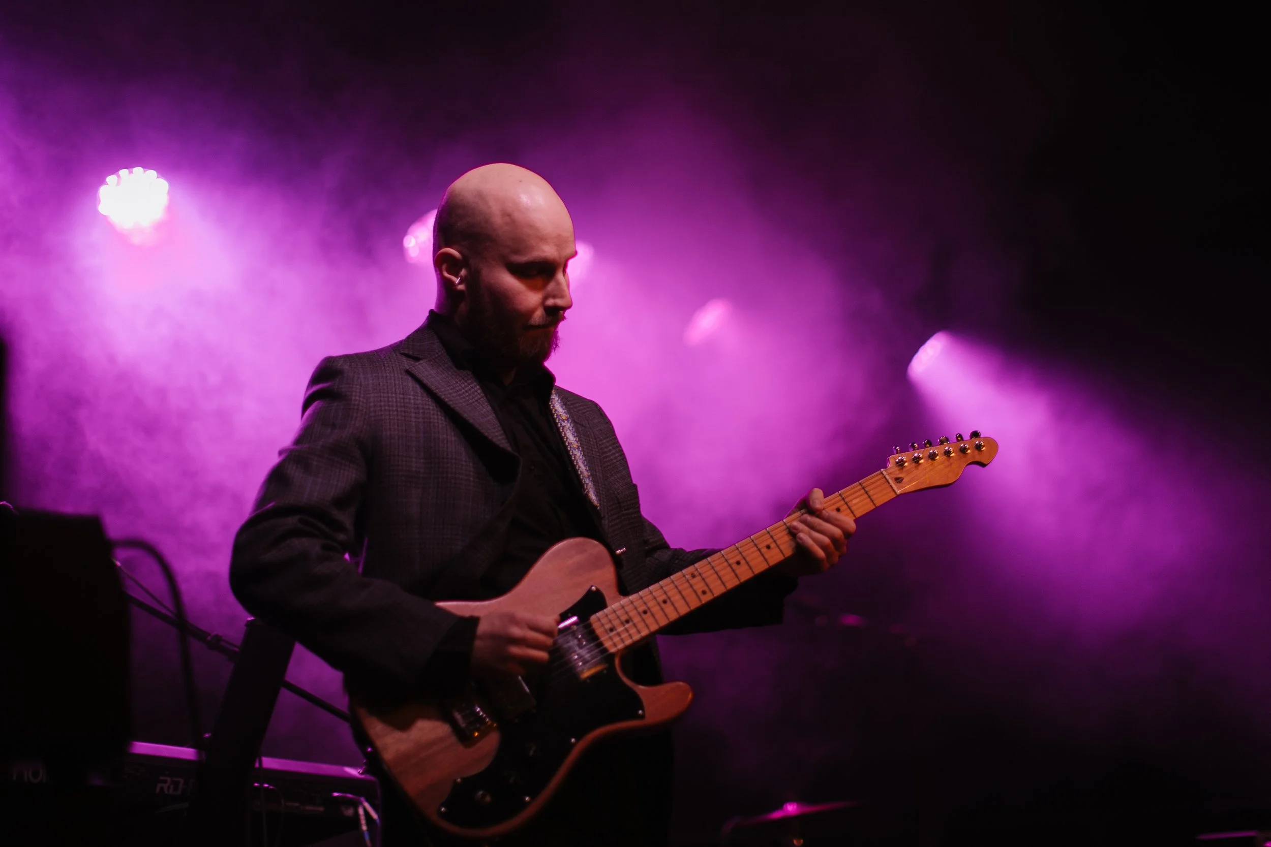 Bald man with a beard playing an electric guitar on stage with purple and pink lighting.