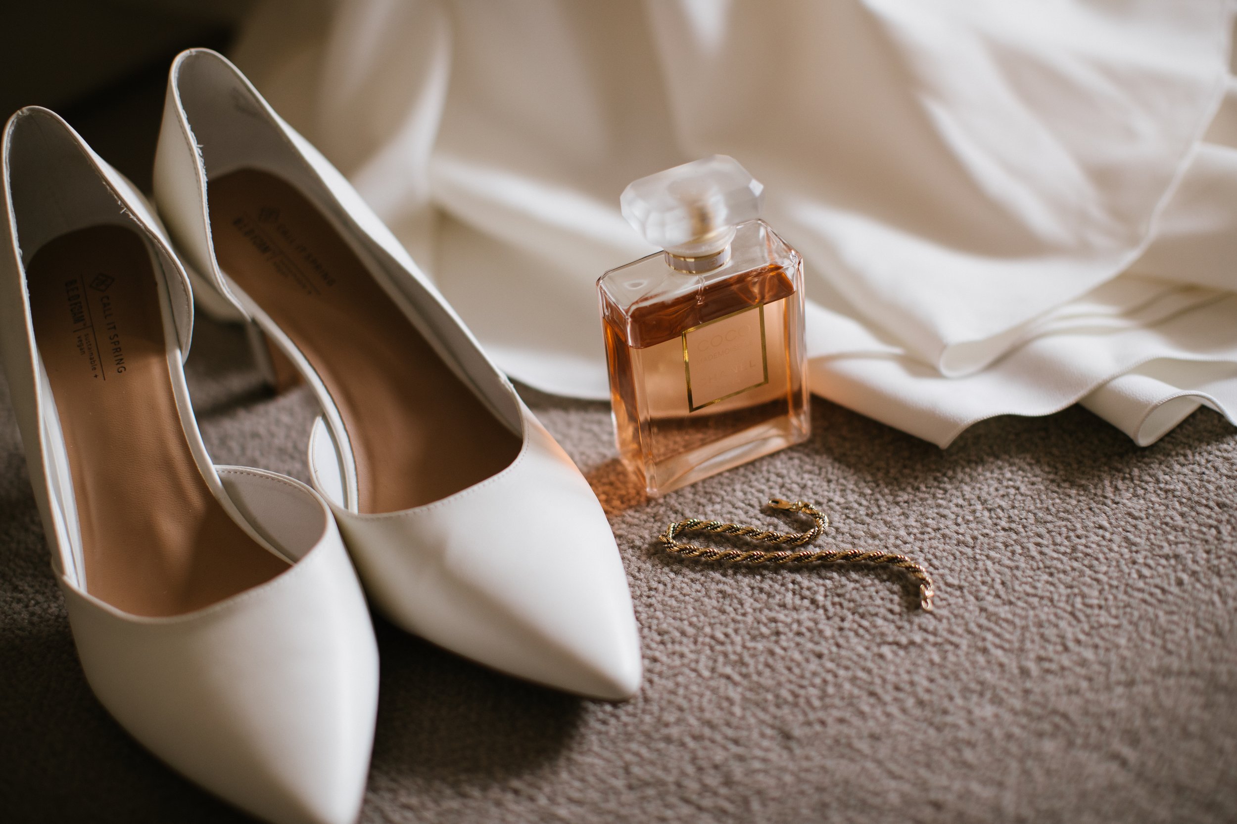 Pair of white high-heeled shoes alongside a bottle of perfume, a gold bracelet, and a ring on a beige carpet with a white fabric backdrop.