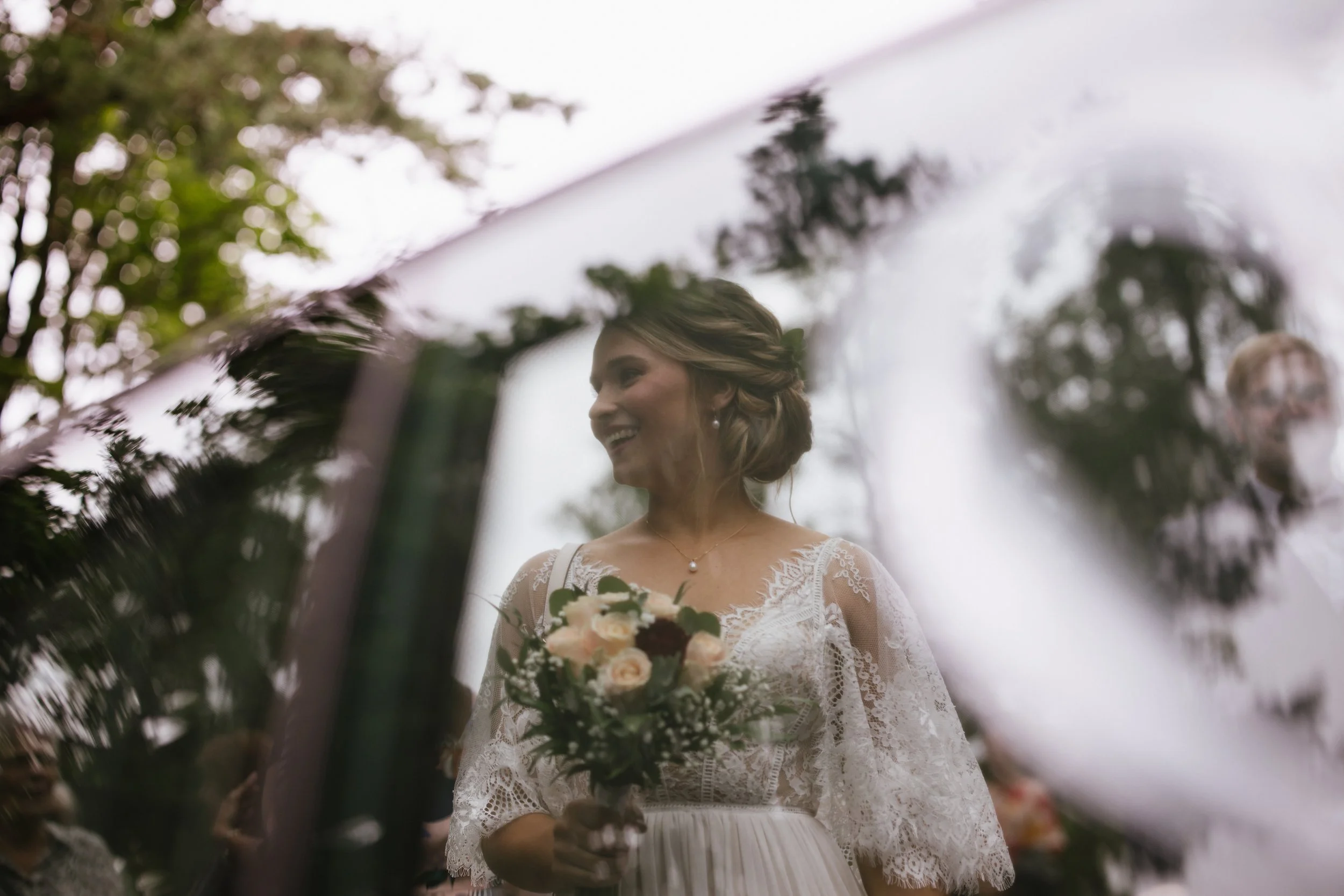 A woman in a white lace wedding dress holding a bouquet of flowers, smiling, as seen through a reflective surface with trees in the background.