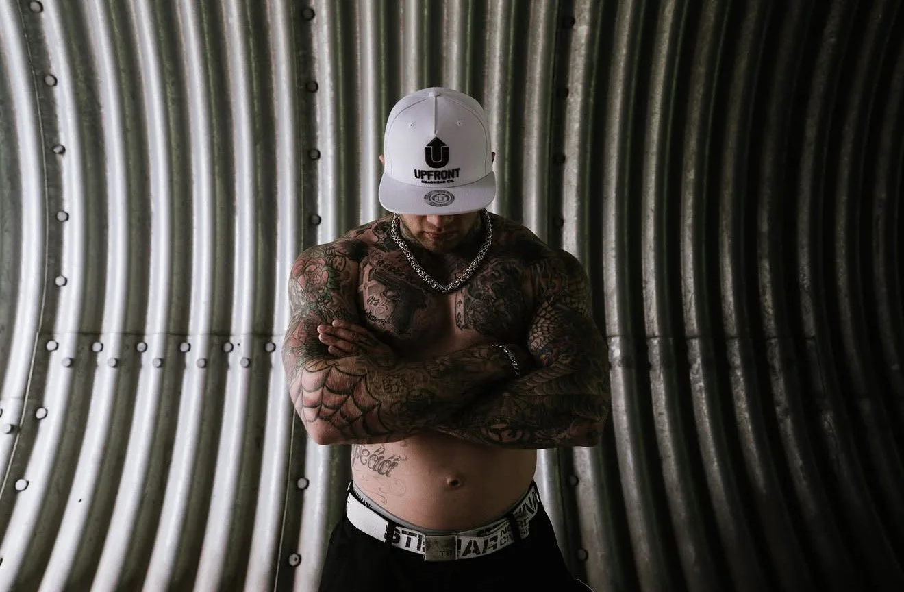 A shirtless man with tattoos on his arms and chest, wearing a white baseball cap with a logo and the word 'UPFRONT', a silver chain necklace, black pants, and a white belt, standing against a corrugated metal wall with arms crossed and head slightly 
