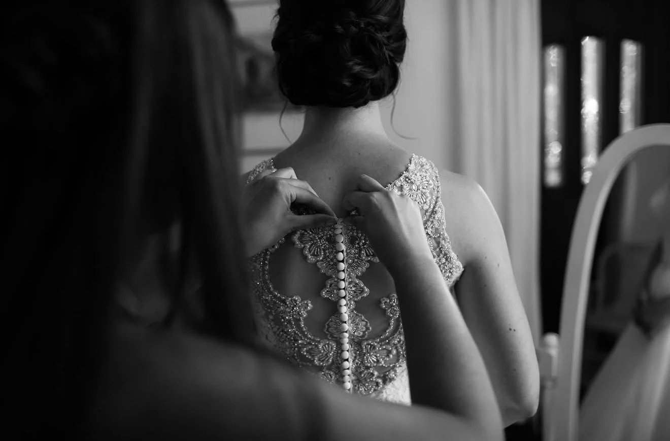 A woman is helping another woman button up a detailed, beaded dress with intricate embroidery and a row of buttons down the back, in a room with natural light coming through windows.