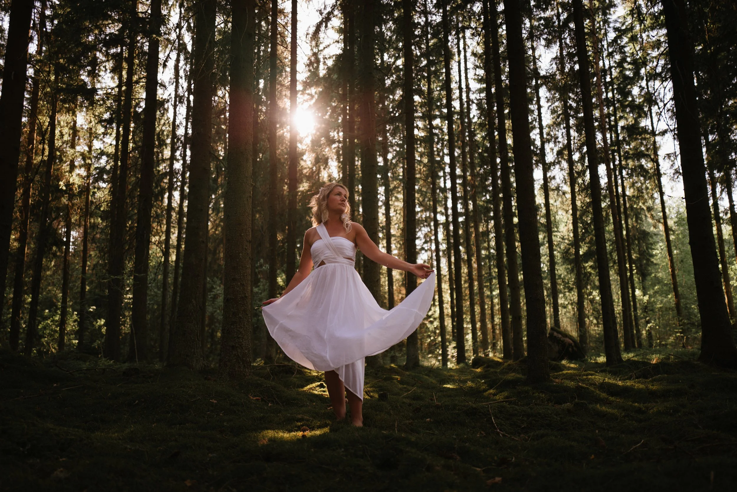 A woman in a white dress standing in a forest during sunset, surrounded by tall trees, with light filtering through the branches.