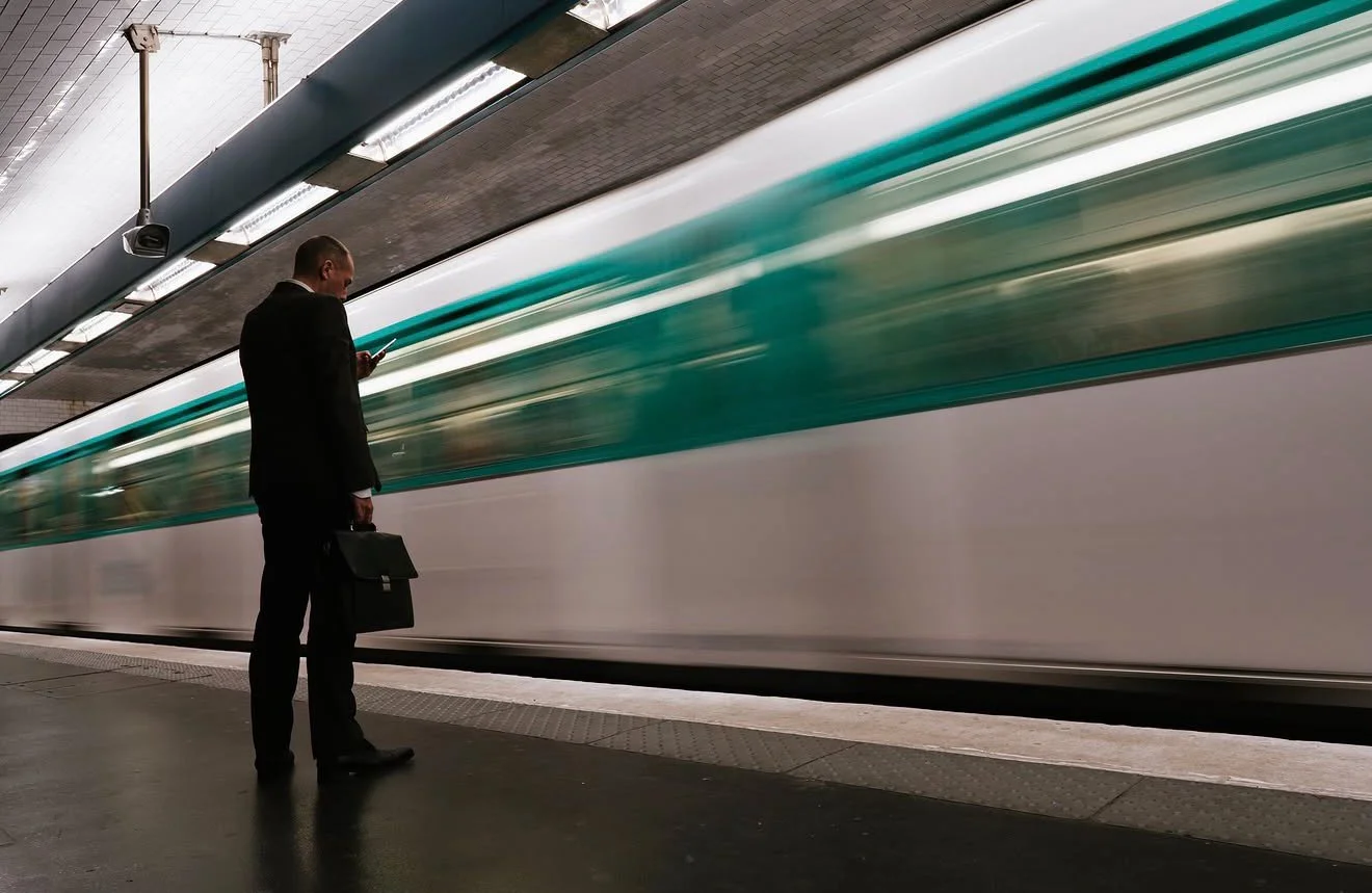 A man in a business suit standing on a subway platform, looking at his phone and holding a briefcase, with a moving train passing by behind him.