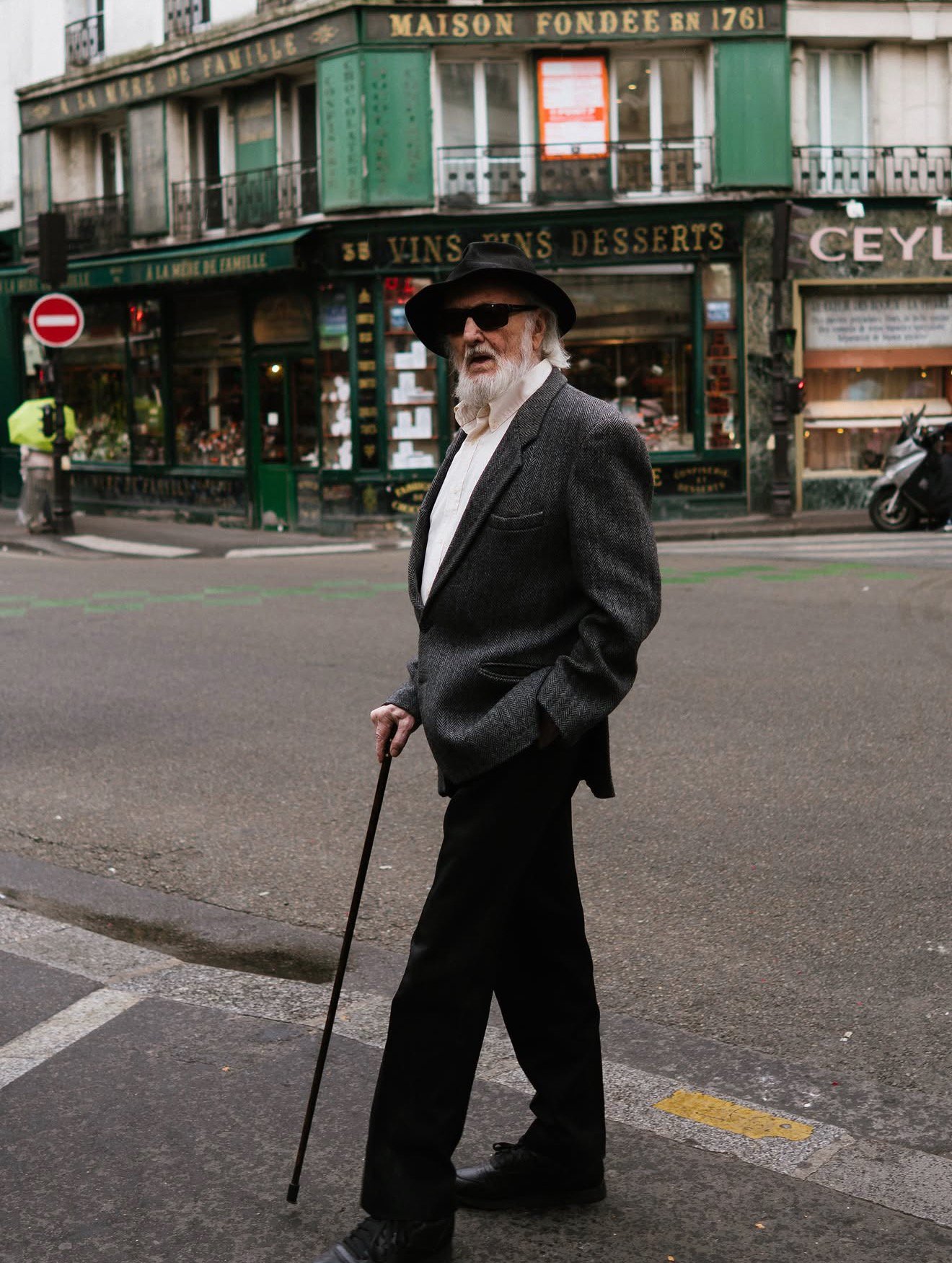 An elderly man with a white beard and wearing sunglasses, a black hat, a gray blazer, and black pants, walks with a cane across a city street in front of a storefront with French signage. The storefront has green and black signage, and there is a mot