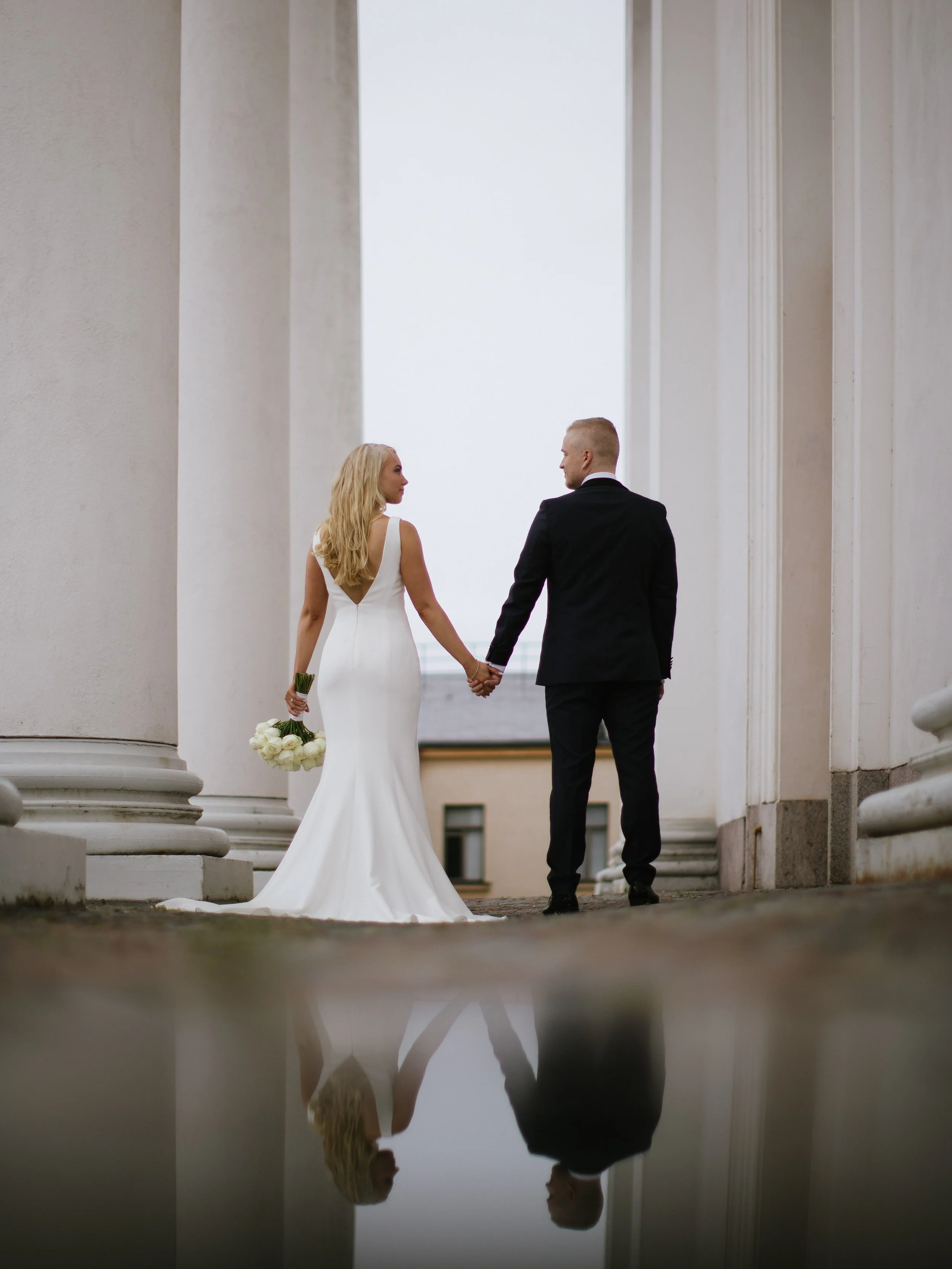 A bride and groom holding hands, walking through a grand, white architectural structure with large columns, as reflected in a shiny floor.