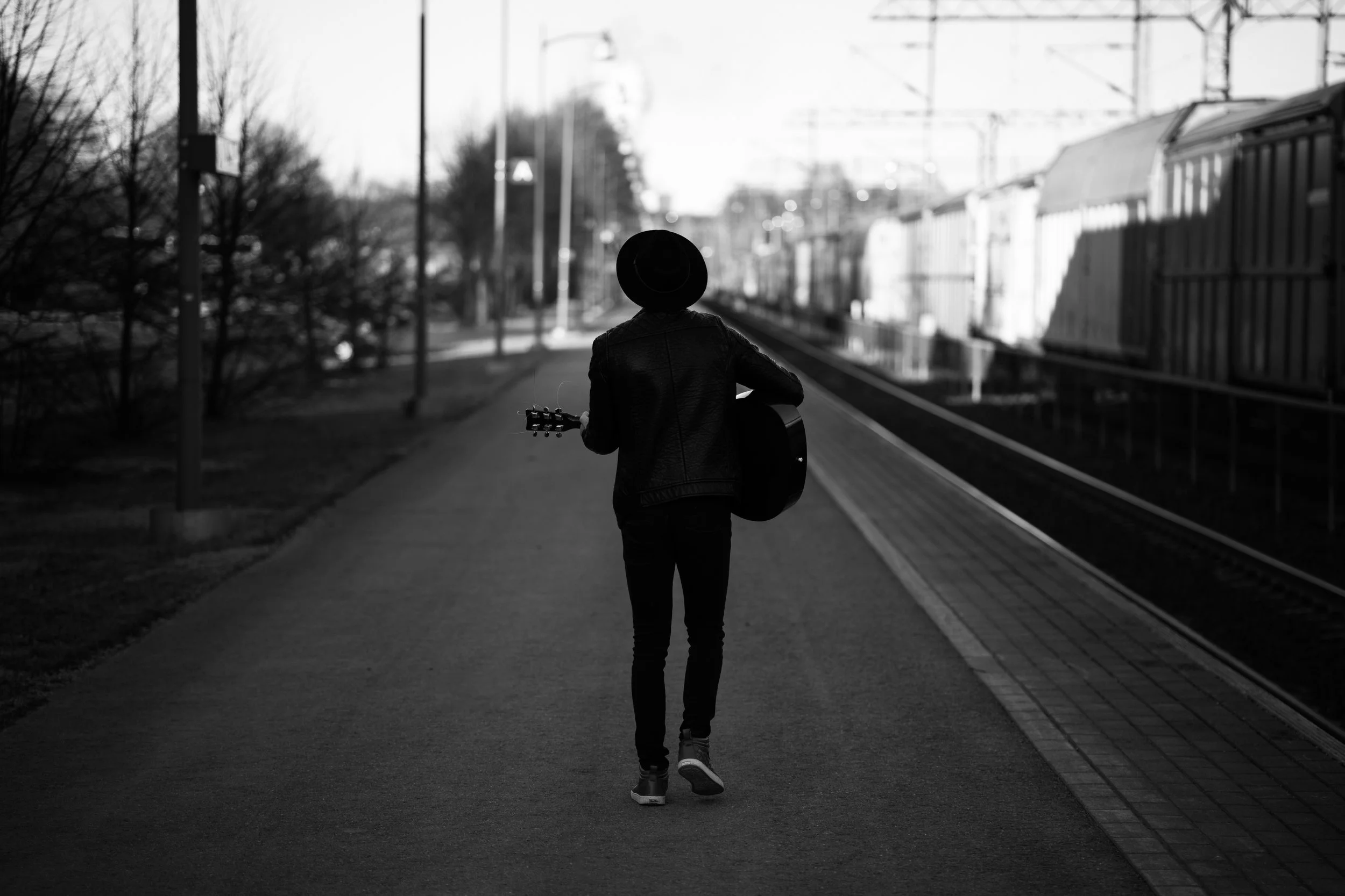 A person walking down a sidewalk holding a guitar, wearing a hat and a leather jacket, with train tracks and train cars on the side, in black and white.