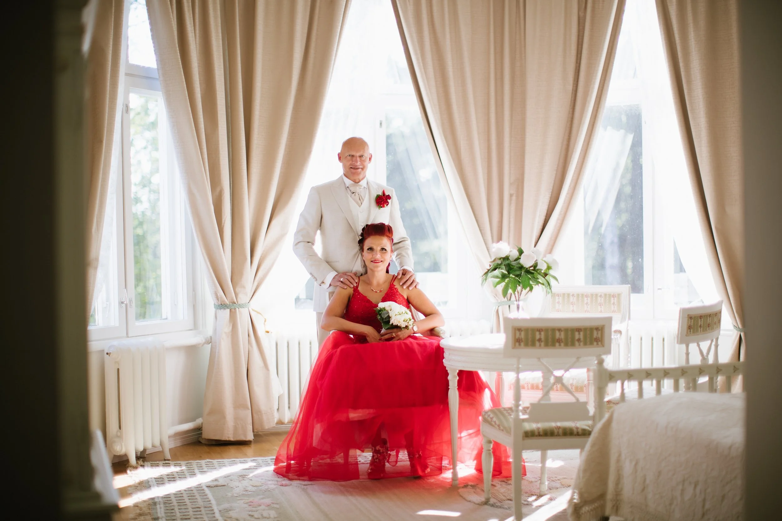 A man in a white tuxedo standing behind a woman in a red dress sitting at a table with a bouquet of white flowers, in a room with large windows and cream-colored curtains.