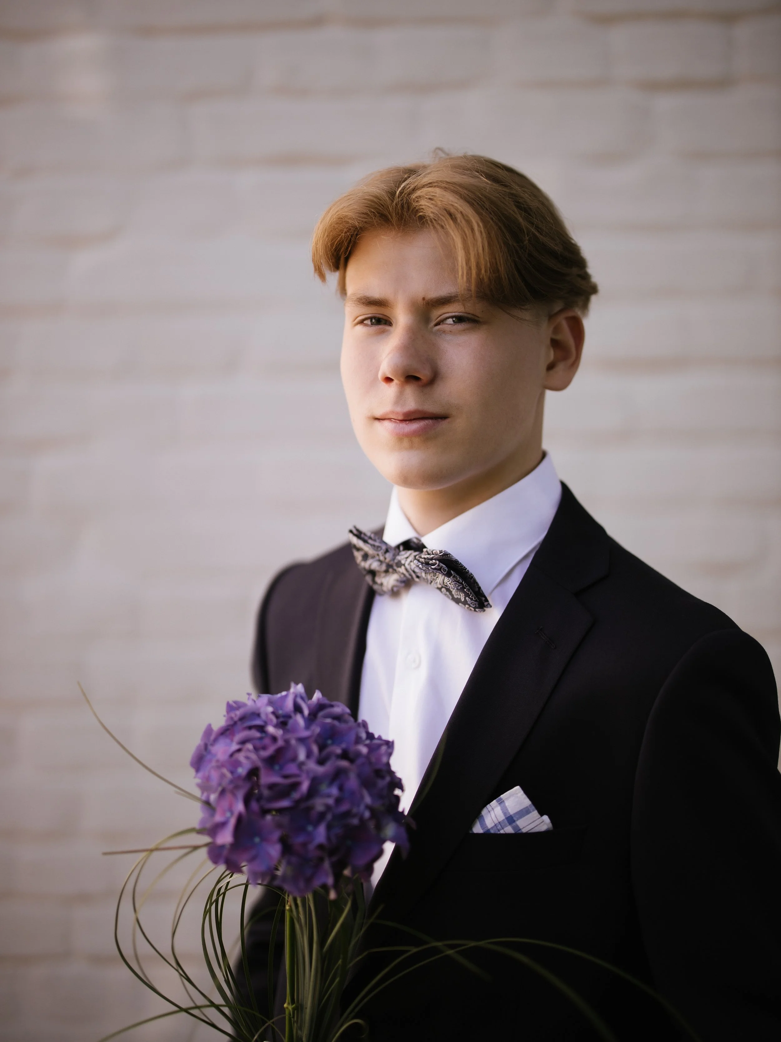 A young man dressed in a tuxedo with a patterned bow tie, holding a bouquet of purple flowers in front of a light-colored brick wall.