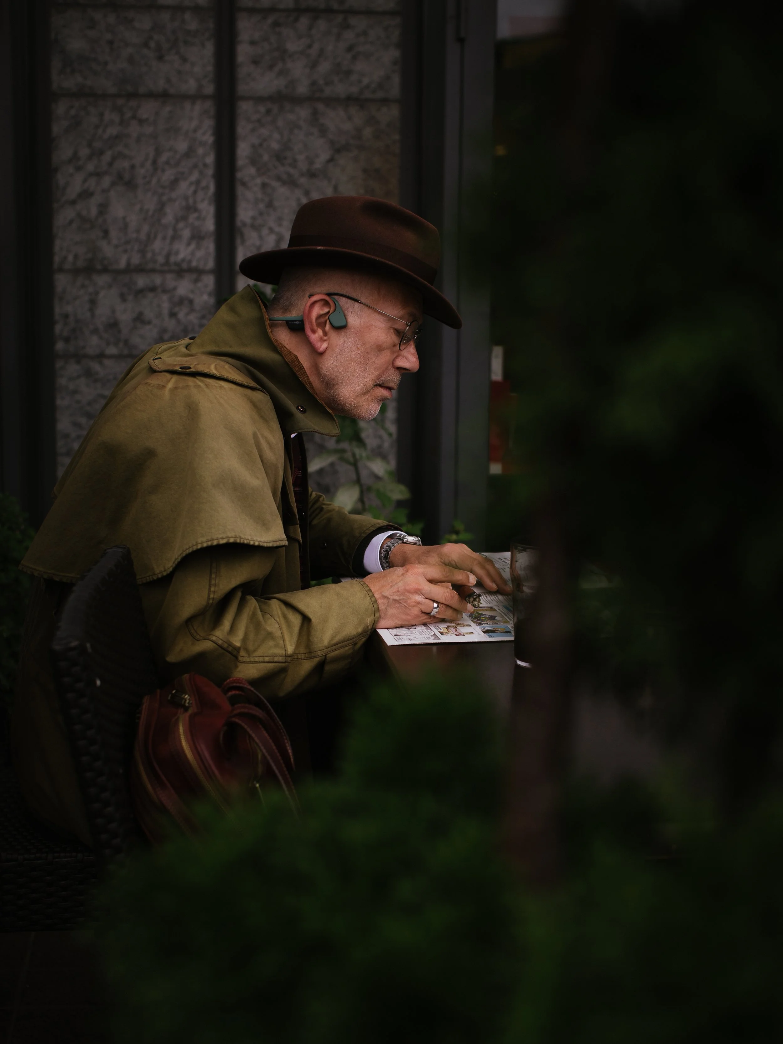A man wearing a brown hat, glasses, and a beige jacket sitting at a table outside, looking down at some papers or a menu.