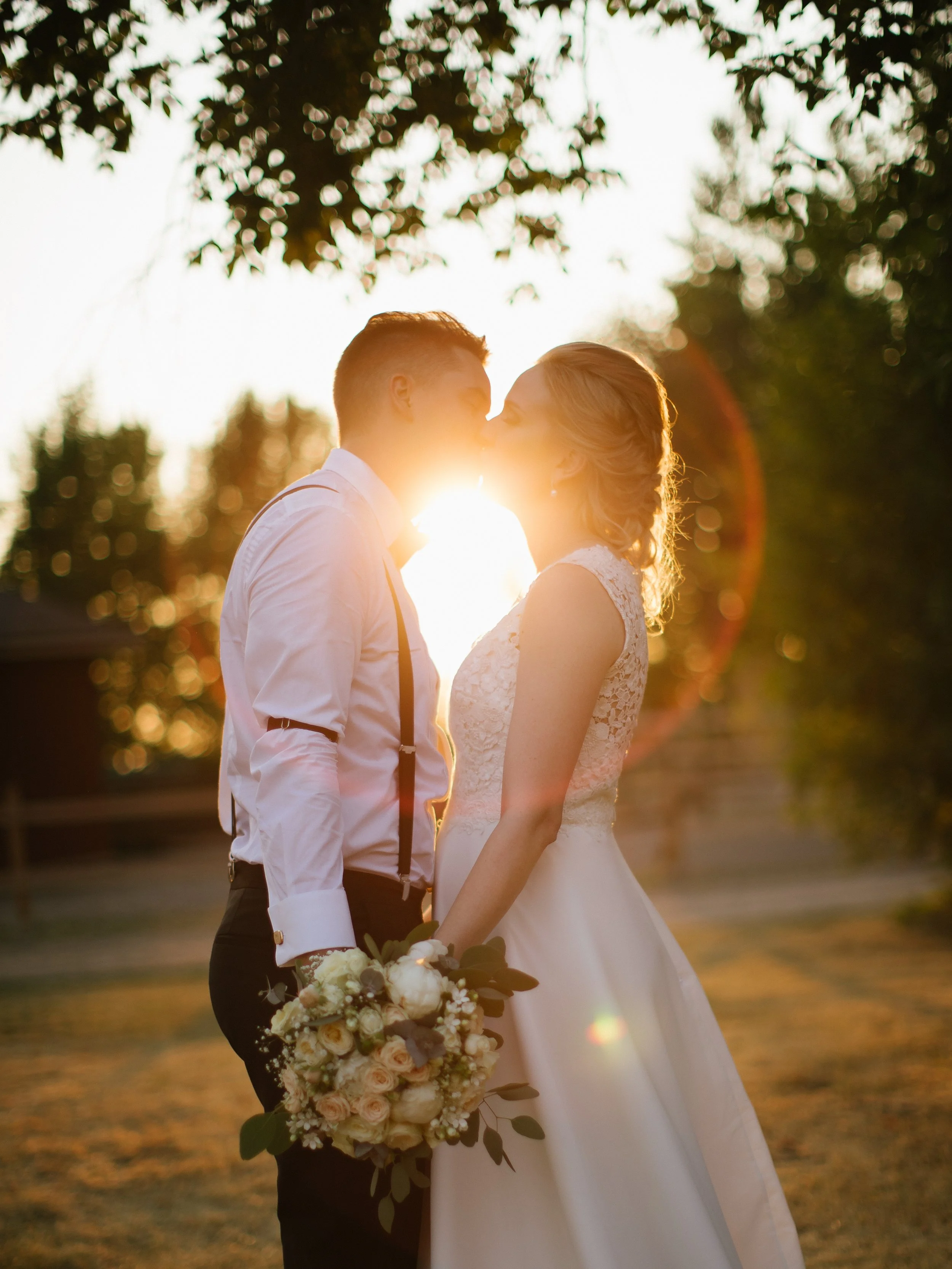 A bride and groom are kissing outdoors during sunset, with the sunlight creating a lens flare and warm glow around them. The bride is wearing a white lace wedding dress and holding a bouquet of flowers, while the groom is dressed in a white shirt wit
