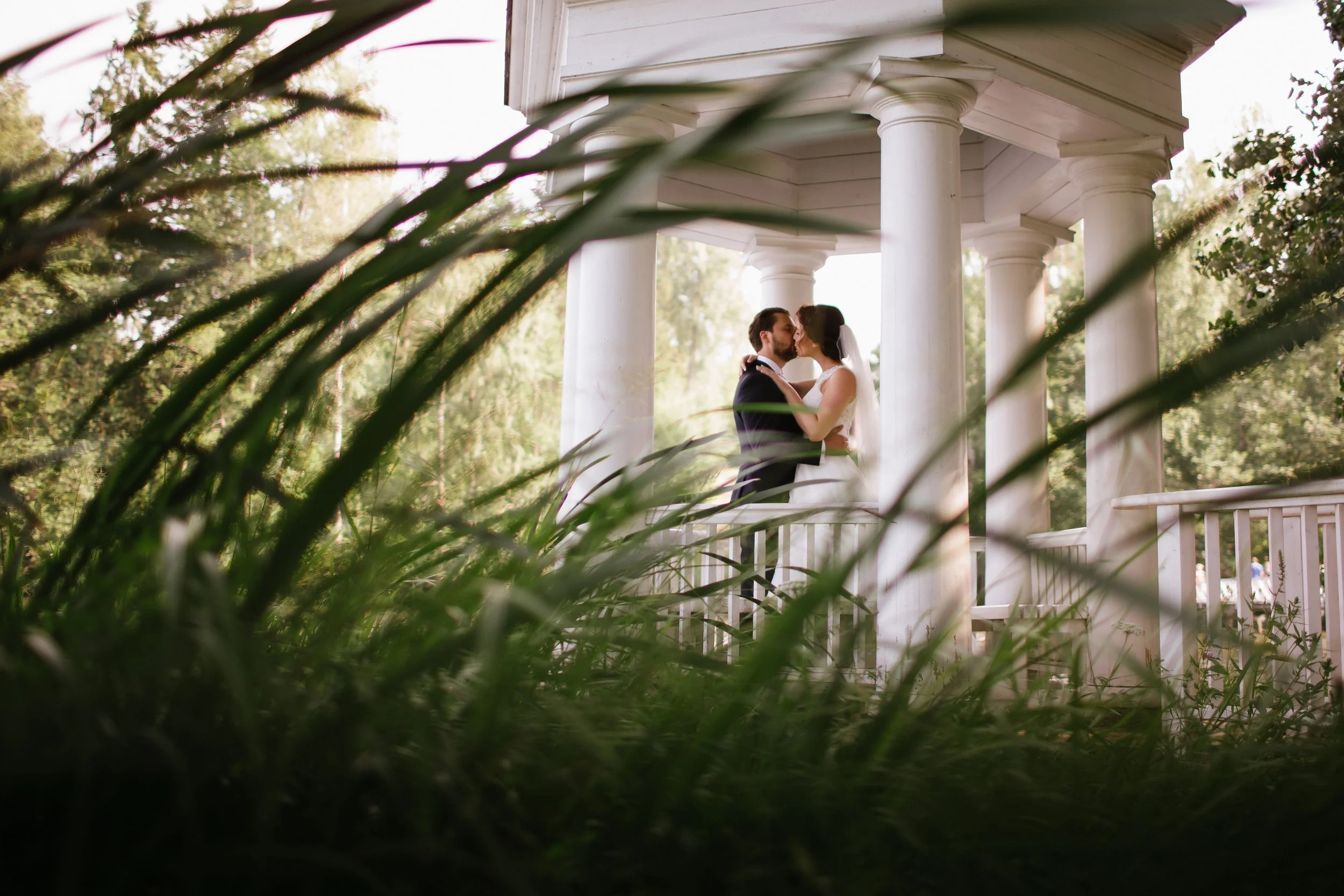 A bride and groom kissing under a white gazebo surrounded by greenery.