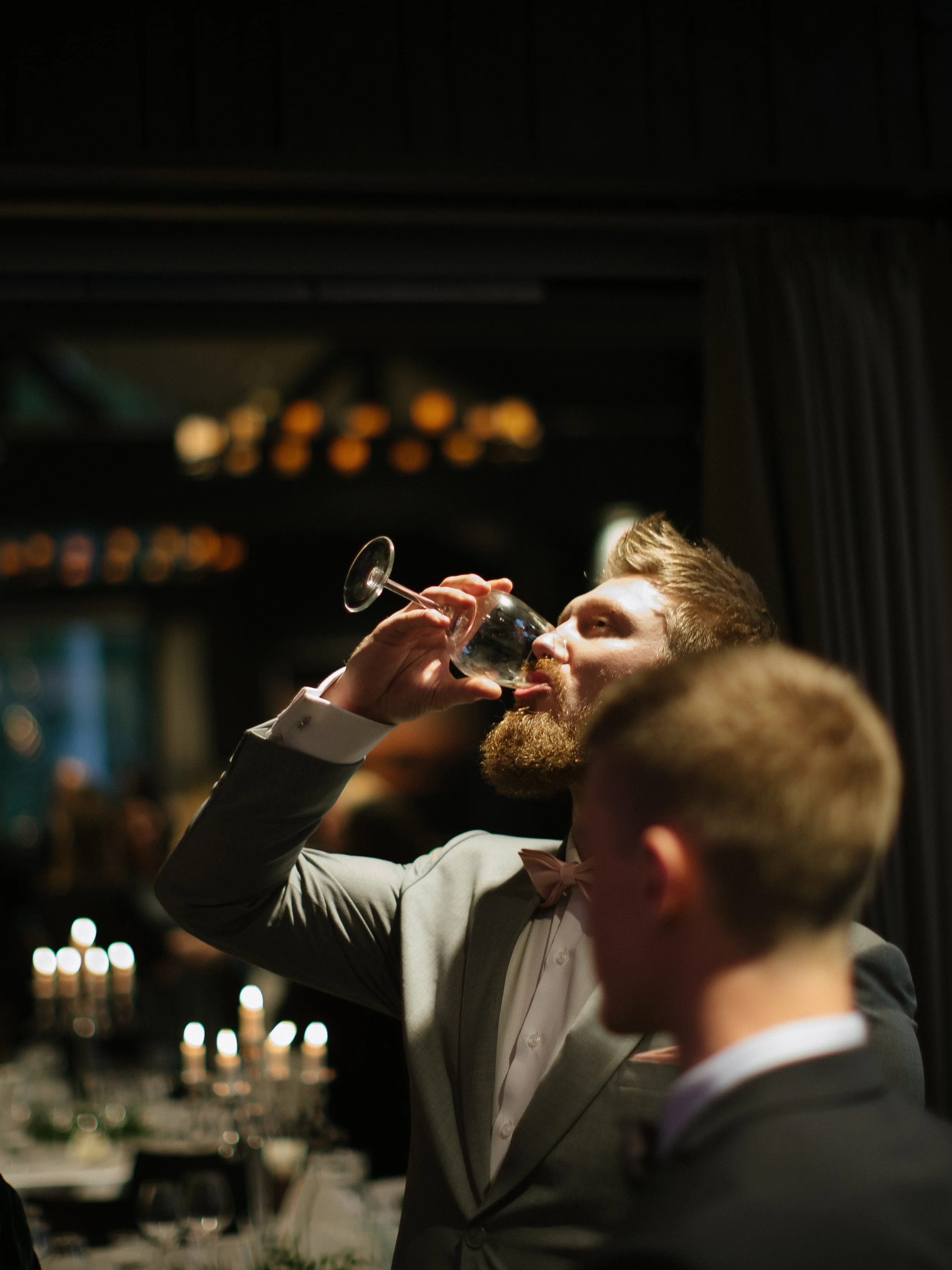 A man with a beard wearing a tuxedo and bow tie is drinking wine at a formal event in a dimly lit room with blurred background of lights and tables.