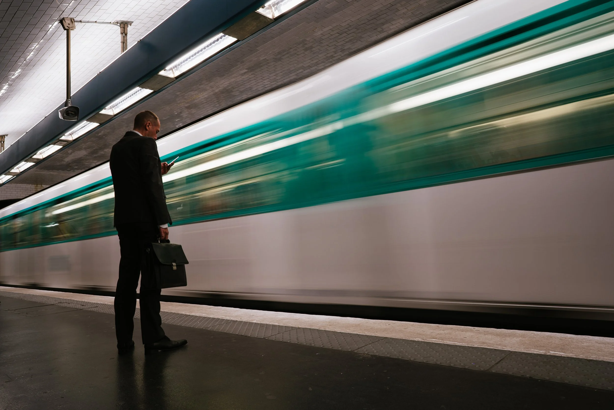 A man in a suit is standing on a subway platform, looking at his phone and holding a briefcase. A fast-moving train passes by behind him in a subway station with tiled walls and overhead lighting.