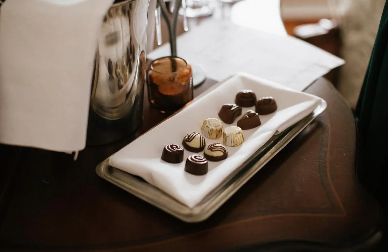 Assorted chocolates on a white cloth on a dark wooden table with a glass container and a metal appliance in the background.