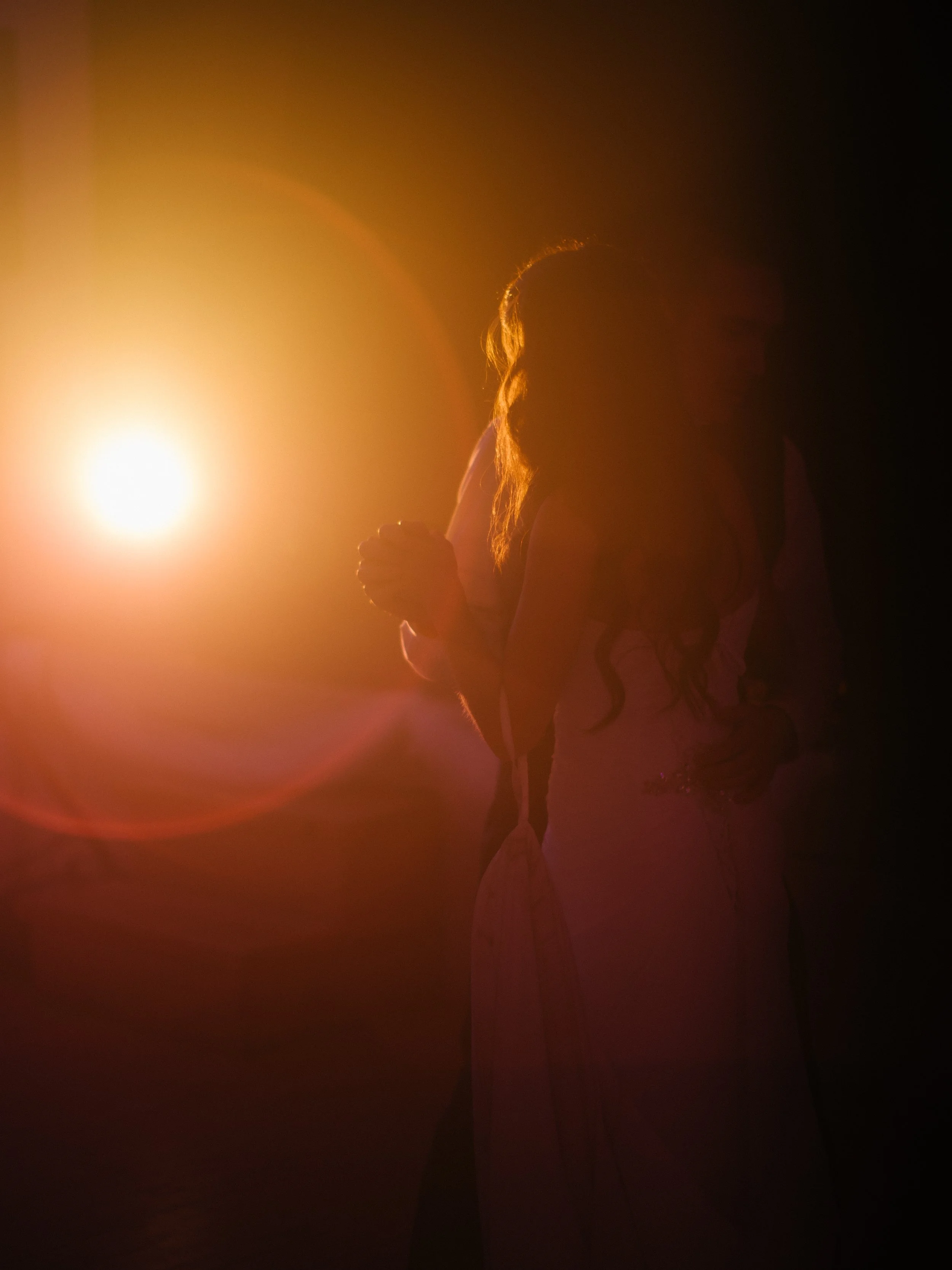 A woman standing against a bright, warm light source in a dark setting, with her hair illuminated by the light and her hands clasped in front of her.